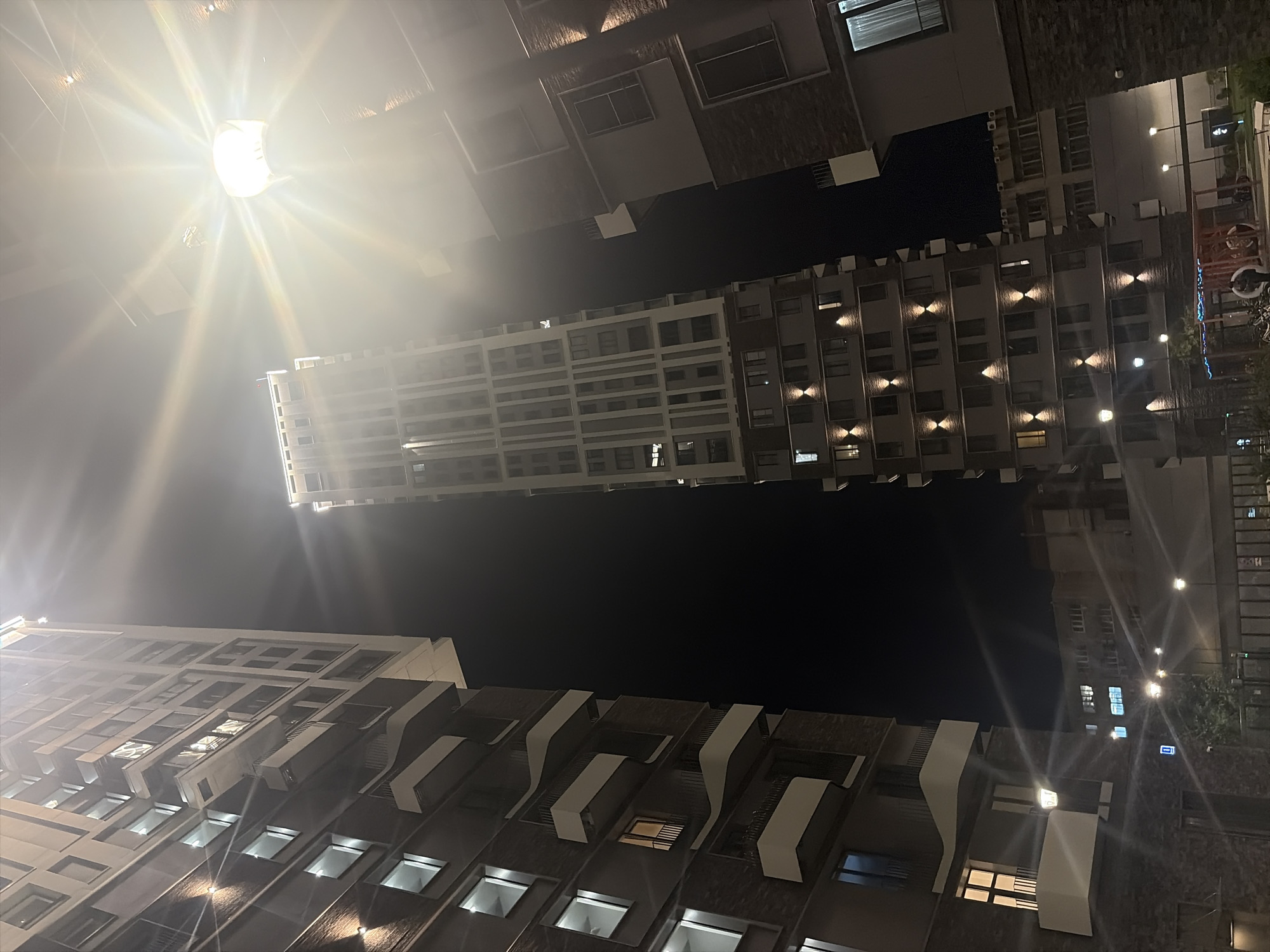 This is a nighttime photograph of a modern residential complex in Moscow, Russia. The image captures several tall buildings with illuminated windows, suggesting occupancy. A prominent streetlamp with a strong glare is positioned in the upper right quadrant, casting light across the scene and creating starburst effects. The buildings feature balconies and distinct architectural elements, with varying facade materials including brick and lighter-colored sections. The sky is dark, indicating it is nighttime. In the foreground, a fence and some greenery are visible. The overall impression is of an urban residential environment at night.
