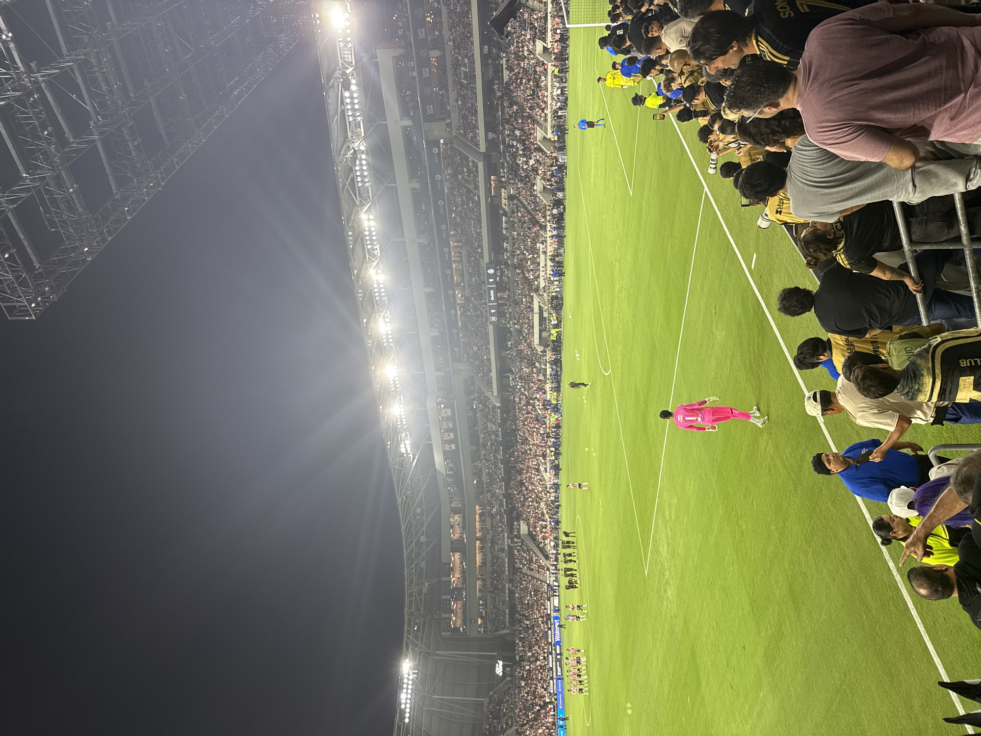 The image depicts a nighttime soccer game in progress at a large, modern stadium, likely in Los Angeles, USA, based on the stadium's architecture and the presence of the "Ford" sign. The field is illuminated by bright stadium lights, and the stands are filled with a diverse crowd of spectators. The perspective is from the stands, looking down onto the field and across to the opposite stands. On the field, two teams of players are visible, one team in red and white striped uniforms and the other in dark uniforms. A goalkeeper in a neon pink jersey stands near the center of the field. Spectators in the foreground are wearing casual attire, with some holding up phones, possibly to take photos or videos. The overall atmosphere appears to be energetic and engaged, with the focus on the sporting event.
