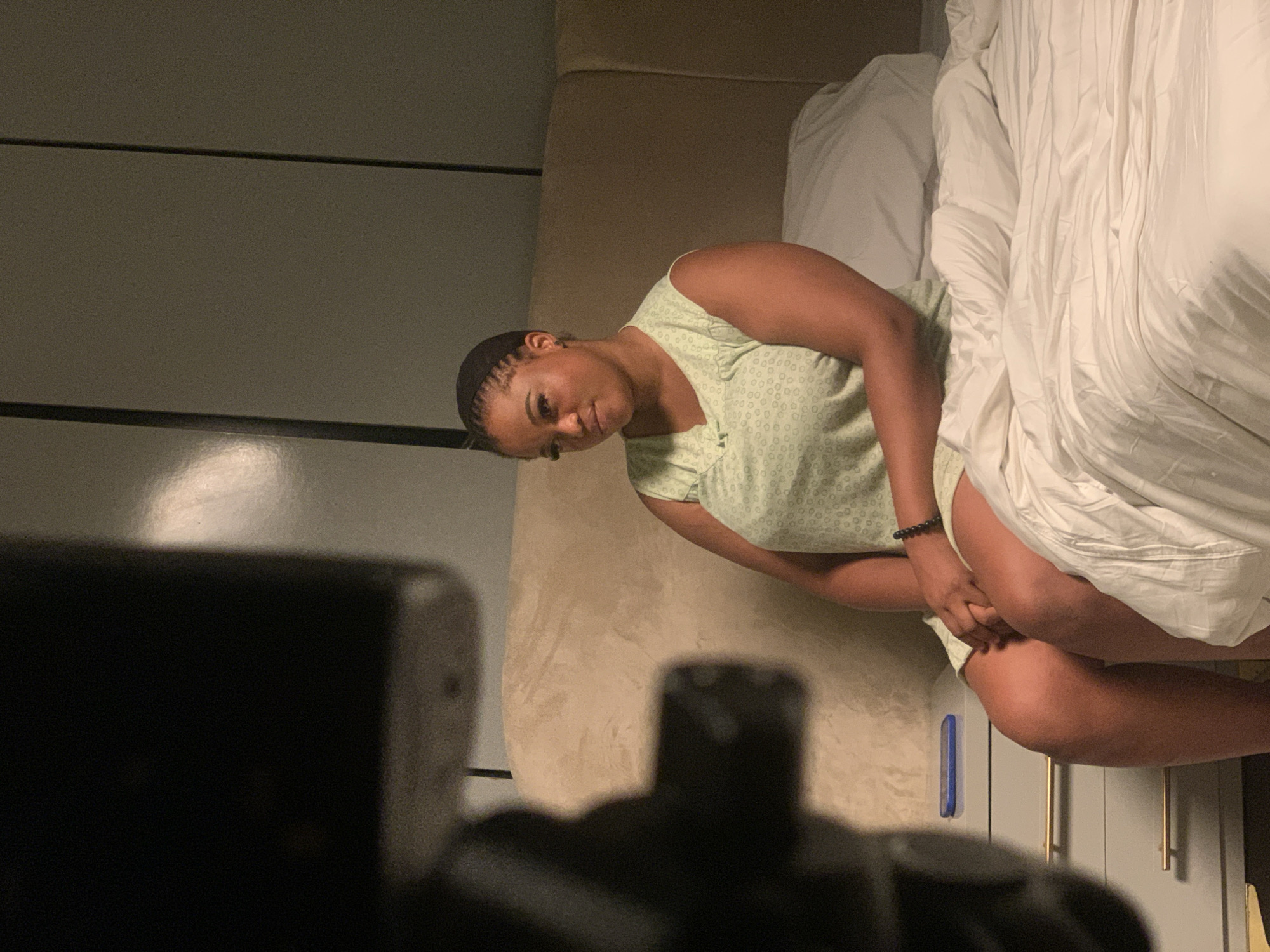 The image displays an indoor scene within a bedroom in Moba, Nigeria. A single adult female is seated on the edge of a bed, positioned slightly to the left of the frame's center. She is observed facing slightly to her left, with her head turned, looking directly towards the viewer. She wears a light green, sleeveless top featuring a patterned design and a V-neckline with a bow detail. A dark beaded bracelet is present on her left wrist, and her hands are clasped in her lap.
The bed is furnished with white bedding, including a duvet and a pillow, against a light brown, padded headboard. To the immediate left of the bed, a light-colored nightstand with two drawers, each equipped with a horizontal gold-colored handle, is visible. A blue smartphone rests horizontally on the top surface of this nightstand. The background wall is a uniform light color, marked by dark vertical lines. A large, dark, out-of-focus object, consistent with photographic or video equipment, partially obstructs the lower left foreground of the image. The environment is dimly lit.
