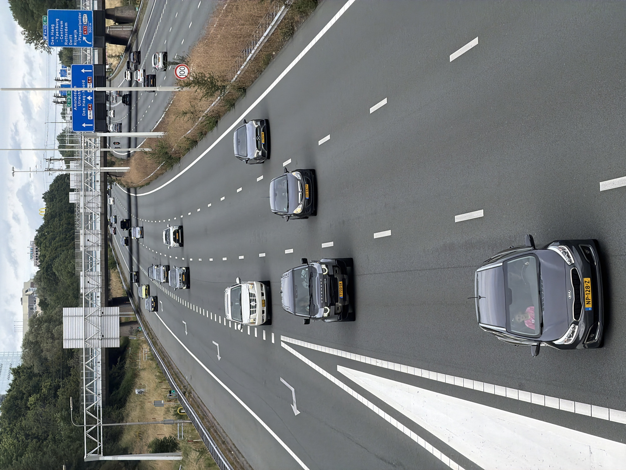 The image shows a view of a multi-lane highway with several cars driving. The highway has white lane markings and a concrete barrier on the right side. Overhead gantries with signs are visible. One sign indicates directions to Amsterdam, Utrecht, Den Haag, and other locations, with the highway designation A4. Another sign indicates directions to Den Haag, Ypenburg, Centrum, Delft, Rijswijk, and Plaspoelpolder, with highway designation A13. A speed limit sign showing "100" is also present. The environment appears to be a mix of highway infrastructure and green vegetation on the side.
In the foreground, a dark gray Kia hatchback with license plate NP-128-Z is driving in the right lane. A young girl wearing a pink top is visible in the passenger seat. Behind this Kia, a dark gray Land Rover Defender with license plate T-720-SK is in the left lane. Further up the road, a white Cadillac Escalade with license plate E-RN5-00 is in the right lane. Other vehicles, including a dark sedan with license plate J-746-TS, are also visible on the highway.
The overall scene depicts a typical traffic situation on a Dutch highway, likely near or within the city of Rijswijk, Netherlands, given the prominent signage. The weather appears to be overcast.