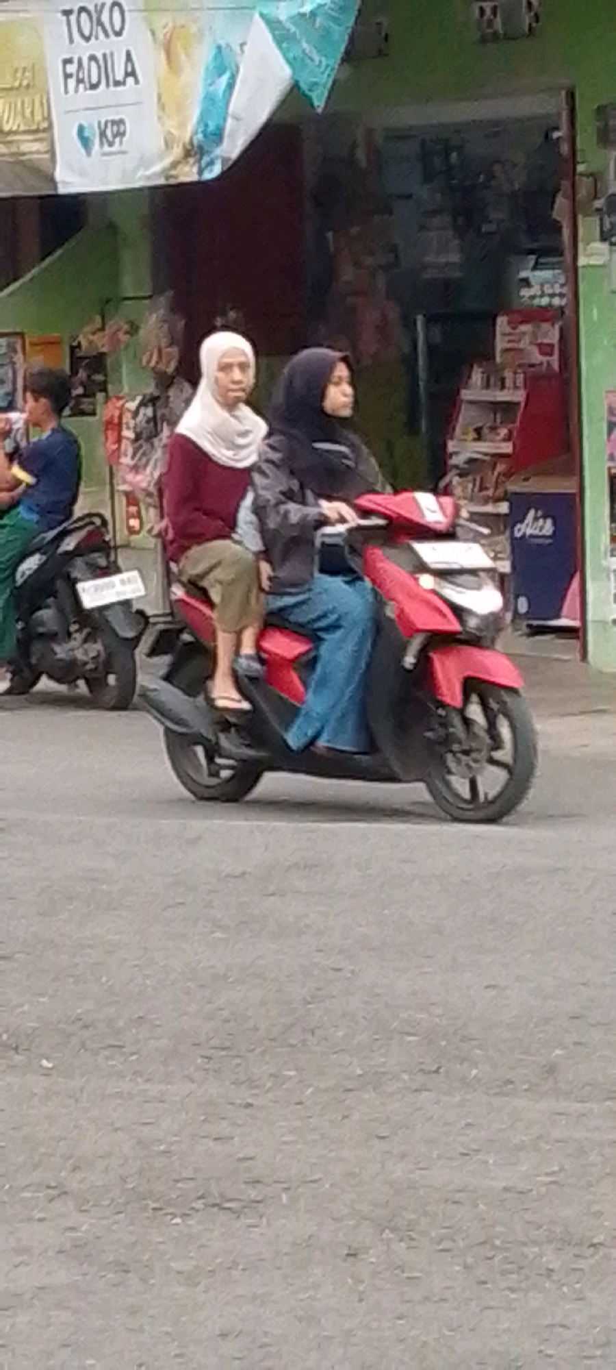 Two individuals, one older with a white hijab and a maroon shirt, and a younger individual with a black hijab and denim jeans, are seated on a red scooter. They appear to be in motion on a paved road. The older individual is in front, and the younger individual is driving the scooter. In the background, there is a building with a green facade and a banner with the text "TOKO FADILA KPP" and a logo that appears to be a heart. The environment suggests a commercial area in Cilaku, Indonesia, with a shop or store visible. Another person on a dark-colored motorcycle is partially visible to the left.