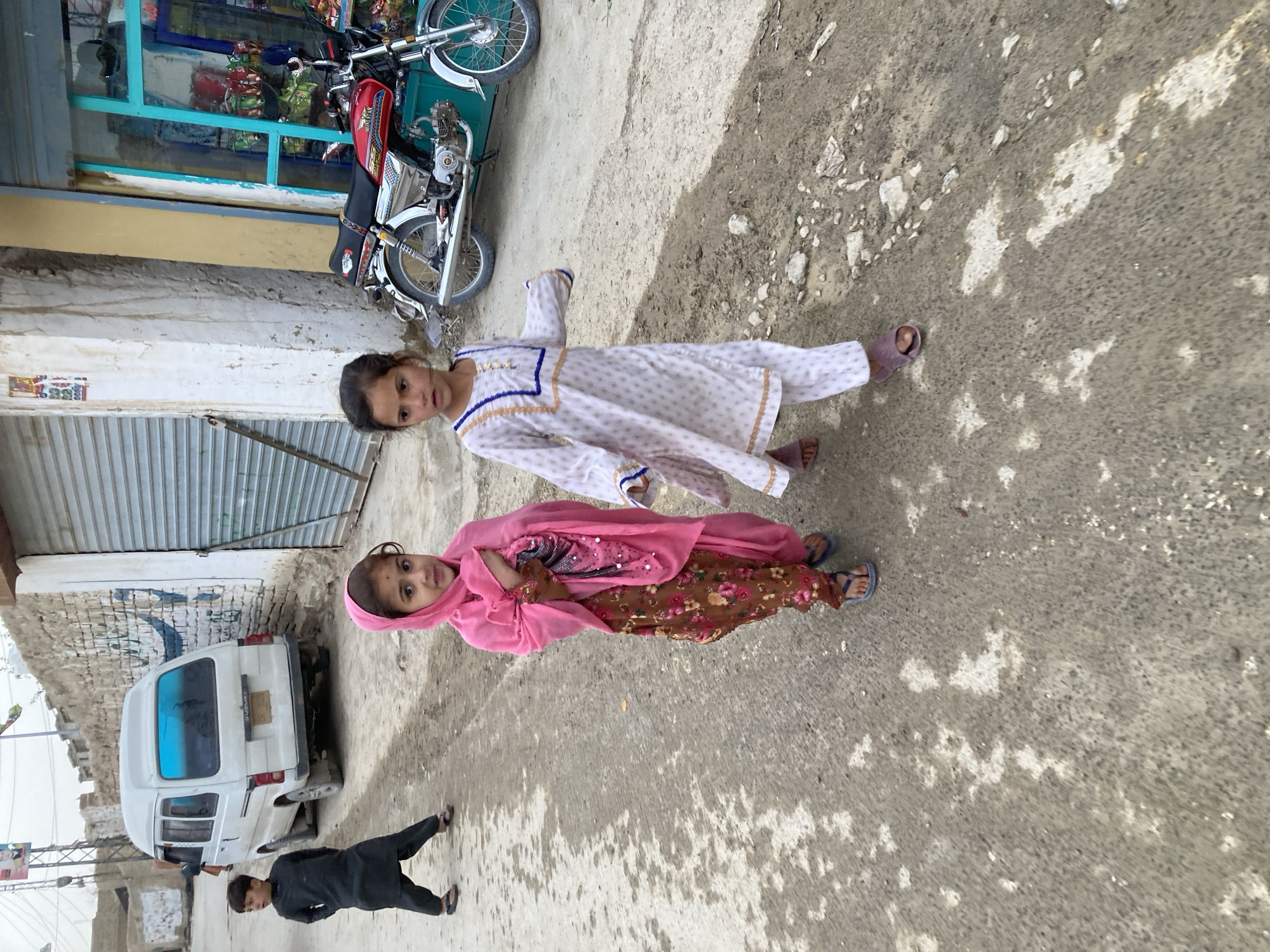 Two young girls and a young boy are on a street in what appears to be Quetta City Tehsil, Pakistan. One girl is wearing a white patterned salwar kameez, while the other is wearing a pink headscarf with a pink and floral patterned outfit. The boy is wearing a black shalwar kameez and walks behind them. A motorcycle is parked to the side of a store with a blue window frame. A white van is parked further down the road. The road surface is cracked and uneven.