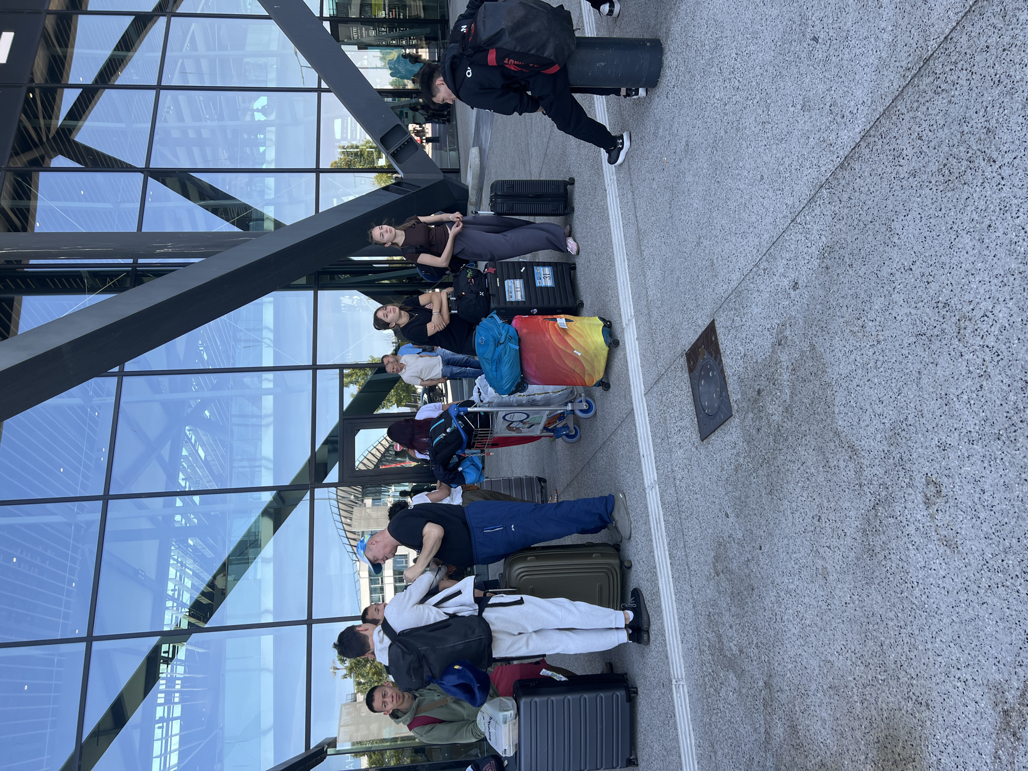 The image depicts an outdoor scene in front of a modern building with a glass facade, likely at the Lyon-Saint Exupéry Airport in Colombier-Saugnieu, France. Several individuals are standing or sitting with luggage. In the foreground, a group of people are gathered, some with suitcases and backpacks. A man in a blue cap and blue pants is interacting with another man in a white tracksuit and backpack. To the right, two young women stand with luggage. Further back, more people and luggage are visible. The architecture of the building features prominent dark grey angular structures and large glass panels reflecting the sky and surrounding environment. The ground is paved with textured grey material. The scene suggests a travel hub, with people preparing for or waiting for transportation.