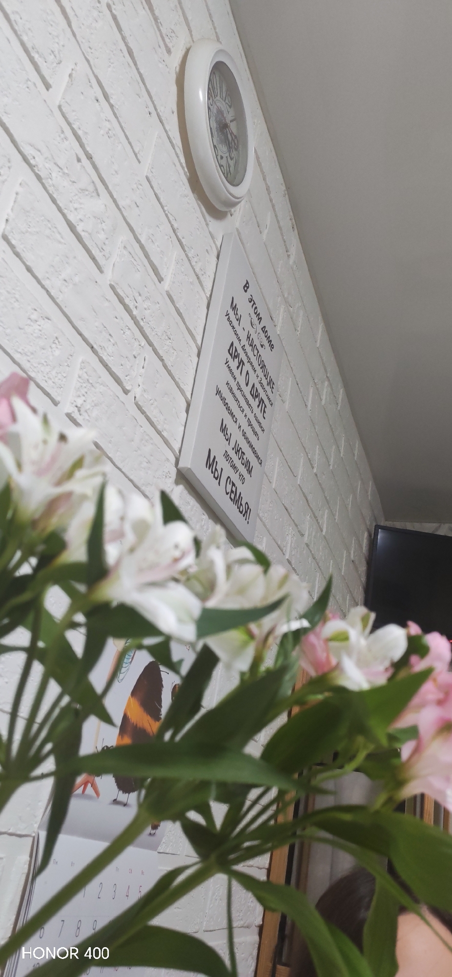 The image shows an interior with a focus on domestic decorations. On a white brick-patterned wall hang a white round clock and a white rectangular plaque with black Cyrillic text, which translates to a family motto, "In this house, we are real...". In the foreground, a blurry bouquet of flowers with white and pink petals and green stems partially obscures a calendar with a butterfly illustration and numerical dates. In the background, a television screen is partially visible, suggesting a living room setting. The text "HONOR 400" is printed at the bottom left, likely indicating the phone model used to take the photo. The overall scene suggests a home interior, likely in Noviy, Russia.