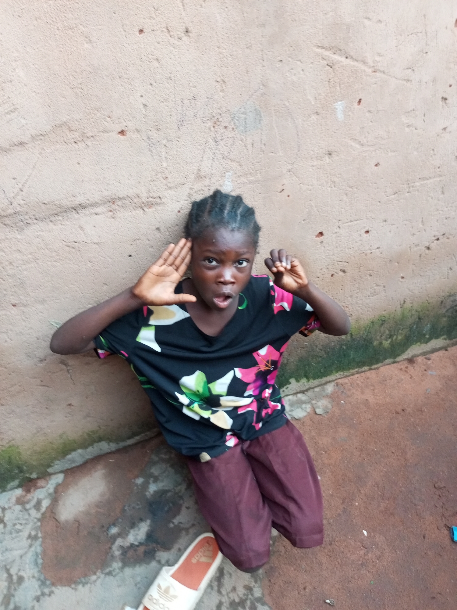 A young Black girl kneels against a rough, textured, light-brown wall, her body angled towards the camera. She wears a black t-shirt with a floral pattern of pink, white, and green flowers, and maroon pants. Her hair is braided into cornrows. Her left hand is raised to her forehead, and her right hand is raised near her face, with her fingers curled. Her mouth is open, as if she is speaking or calling out. The ground beneath her is a mix of reddish-brown dirt and concrete, with patches of green moss or algae. A white sandal with an orange sole and Adidas branding is visible on the ground to the lower left of the frame. The background wall shows signs of wear and tear, with some visible cracks and discolorations. The lighting suggests it is daytime. The image appears to have been taken outdoors in an urban or semi-urban environment.