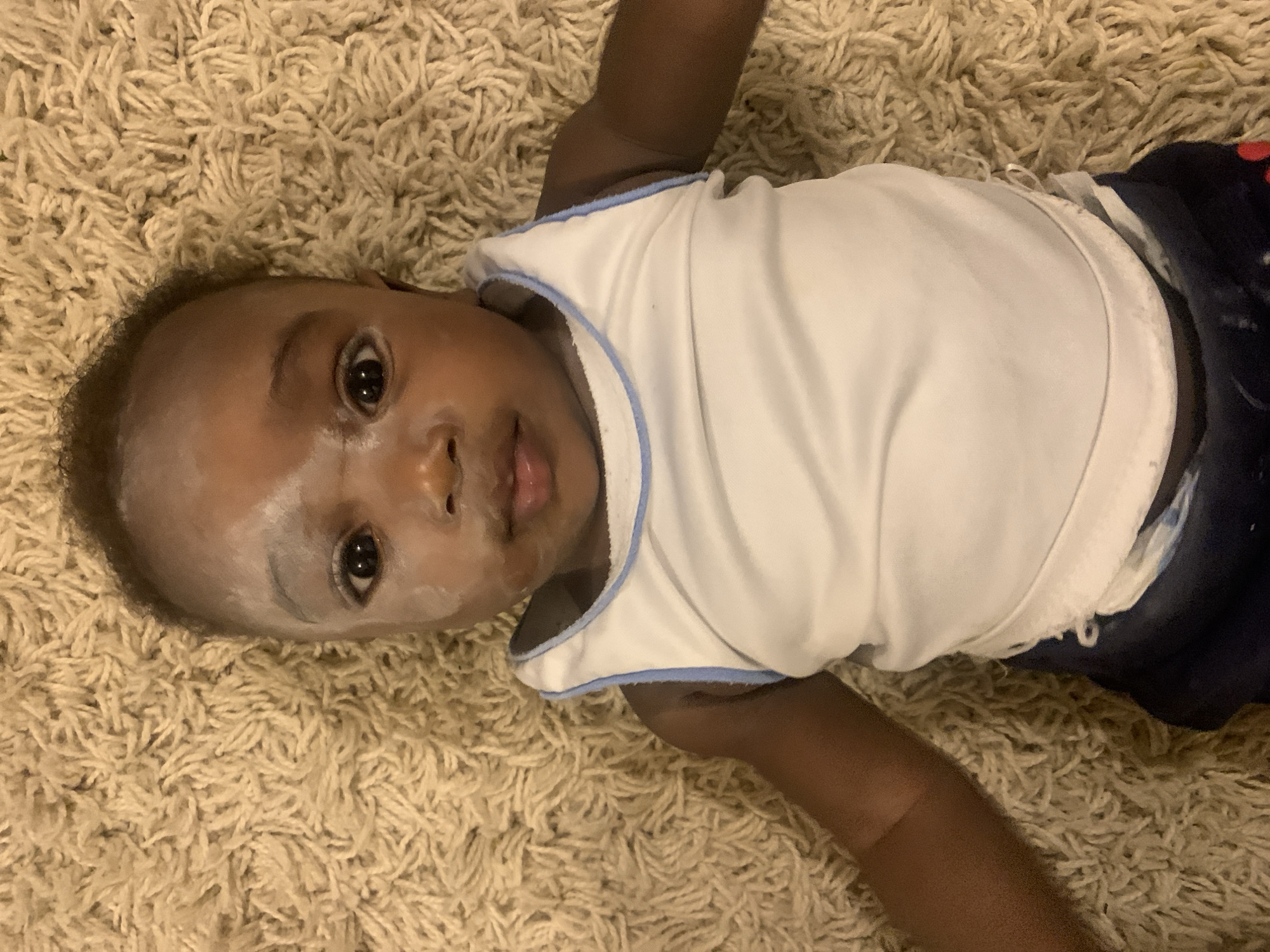 A baby with dark skin lies on a light tan shag carpet. The baby wears a white tank top with blue trim and dark blue shorts. There appears to be a white powdery substance on the baby's face. The baby is looking at the camera with an open mouth. The location is unknown and there is no basis for stating it is in Barakin Akawo, Nigeria.
