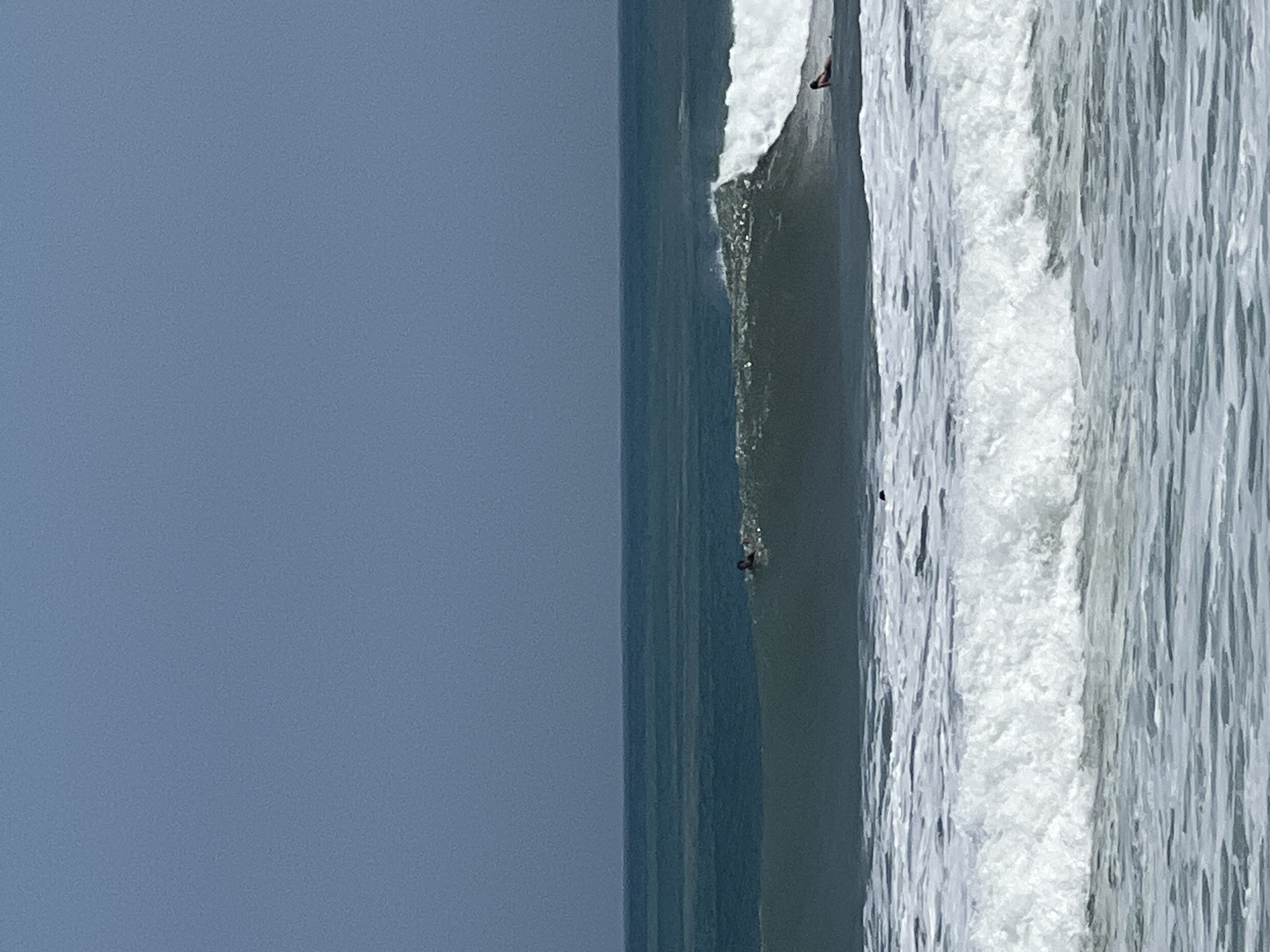 The image depicts a serene ocean scene with a clear, light blue sky. In the mid-ground, a gentle wave is breaking, with white foam cresting. Two surfers are visible in the water. One surfer, wearing dark attire, is paddling on their board in the trough of the wave. Further to the right, another surfer, also on a board, is positioned on the shoulder of the breaking wave, appearing to be in a prone position. The foreground is dominated by the texture of the water, with smaller ripples and waves. The overall impression is one of a calm day at Kuta Beach, Bali, Indonesia, with surfers enjoying the modest surf conditions.