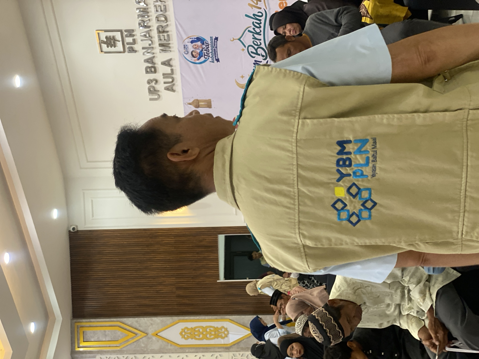 The image shows a man wearing a tan vest with a logo that reads "YBM PLN Yayasan Baitul Maal". He is standing in an indoor setting, possibly an auditorium or meeting room. In the background, there are people seated, some wearing Islamic attire. A banner with text "Tebar kebahagiaan" and "Berkah 1448 H" is visible. The wall behind the banner has text that appears to be "UP3 BANJARMA AULA MERDEK". This suggests the location is in Banjarmasin, Indonesia, at an UP3 (Unit Pelayanan Pelanggan) facility, specifically in the Merdeka Hall. The lighting indicates an artificial indoor environment.