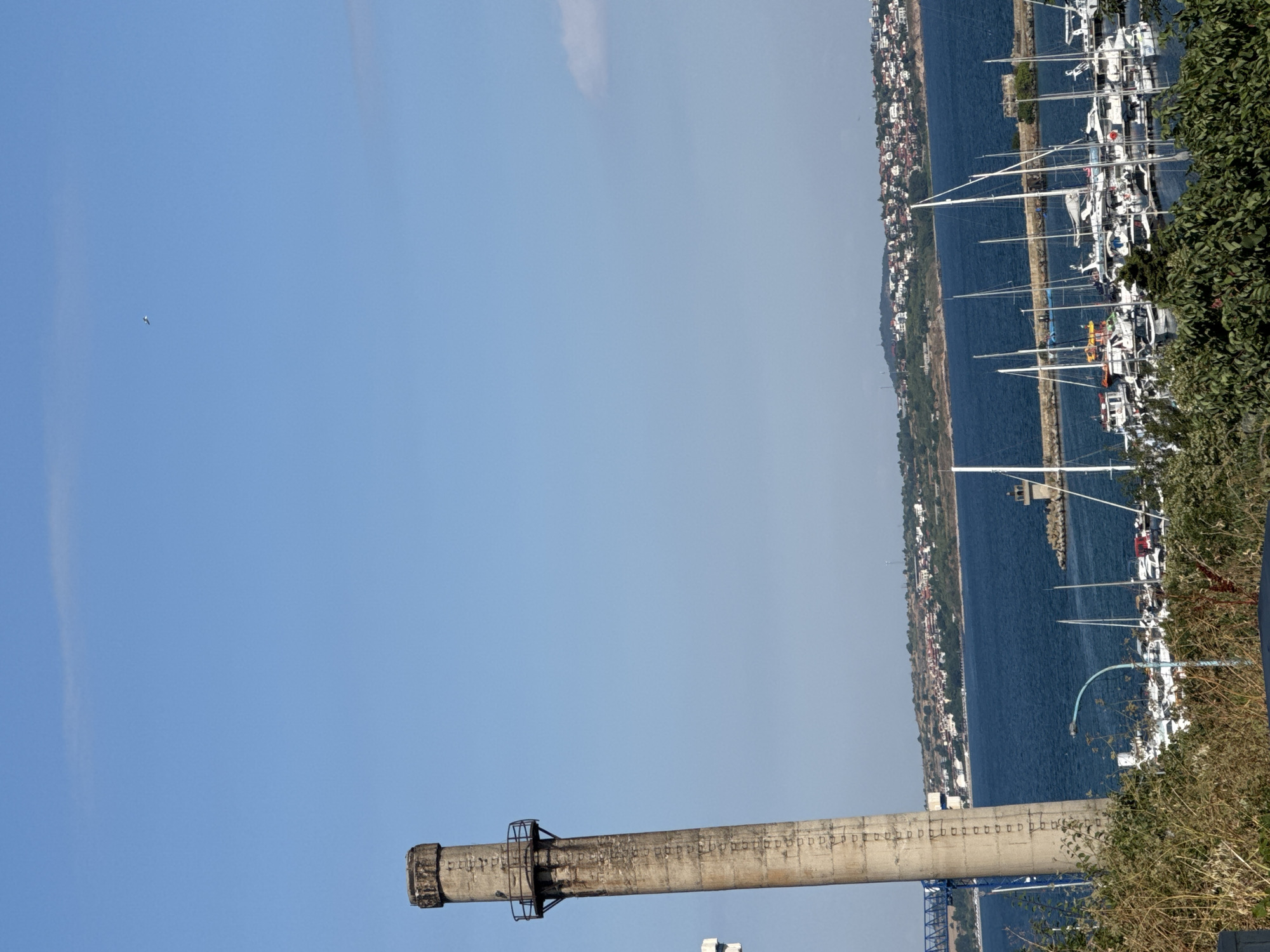 This is a medium-angle, full-frame, vertical shot of a seaside scene in Sozopol, Bulgaria, under a clear, bright blue sky with a few faint clouds and a small bird visible in the distance. In the foreground, a tall, weathered concrete chimney stands on the left, leaning slightly. The chimney has a circular platform with railings about a quarter of the way down its length and appears to have markings or inscriptions running vertically along its surface. Dense green foliage and bushes are situated in the immediate foreground, partially obscuring the base of the chimney.
Beyond the chimney, a body of deep blue water, likely the Black Sea, stretches across the middle ground. A marina is filled with numerous white sailboats of various sizes, their masts creating a striking pattern against the water. A small lighthouse structure is visible on a stone jetty in the middle distance. Across the water, the town of Sozopol is spread out on a gentle slope, characterized by clusters of red-tiled roofs of houses and buildings. The shoreline is lined with more vegetation and some scattered structures. The overall atmosphere is sunny and tranquil, depicting a typical day in a coastal town. There are no people clearly visible in the image.