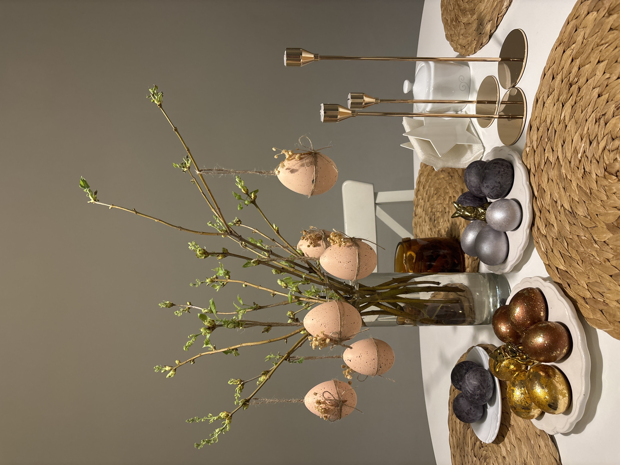 A tabletop display features spring decor, potentially in a home in Chaikovsky, Russia. A clear glass vase holds branches with budding leaves and decorative, light pink Easter eggs speckled with brown and tied with twine. Nearby are three small white plates. One contains dark purple painted eggs, another holds silver painted eggs, and the third plate features gold and bronze painted eggs. A white napkin holder and a small white lidded pot are present, alongside three gold-colored candlestick holders. The display rests on a white surface, with several round woven placemats placed underneath. The background is a plain gray wall.