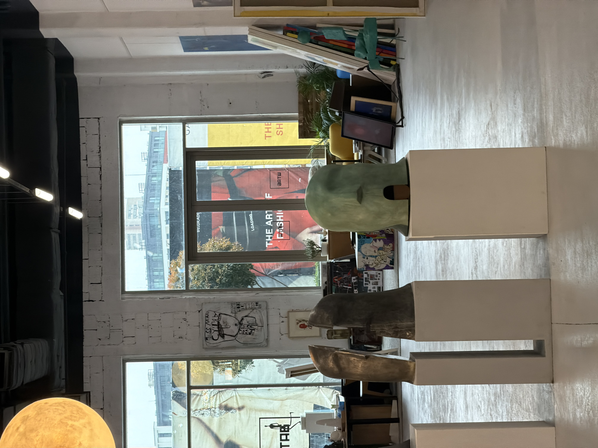 The scene depicts the interior of a contemporary art gallery or studio, characterized by an industrial aesthetic, located in Belgrade, Serbia. The primary visual focus is on three abstract sculptural busts displayed on white rectangular pedestals in the foreground. From left to right, the first sculpture is a metallic, golden-bronze abstract form, the second is a darker, grey-bronze abstract form, and the third is a teal or mint-green, helmet-like head sculpture with a small dark opening at its base.
The interior features white-painted brick walls and a black ceiling with exposed ductwork and linear fluorescent lighting fixtures. Large windows dominate the background, allowing natural light to enter and revealing an urban landscape of buildings and trees outside. One of the window panes on the right displays a red banner with partially visible text, including "THE ART OF" and "BTW," and another area clearly shows "BELGRADE," confirming the city.
Between the two main windows, a large, rectangular, abstract painting with black markings and Cyrillic text ("Јес мао Сутра") on a white background hangs on the wall. Below it, a smaller framed artwork is visible. To the left, various art objects, including a white sculptural figure on a stand, are situated near a wooden table. To the right, an assortment of framed artworks, a yellow armchair, green potted plants, and a light-blue sculptural chair are arranged against the wall and on the floor. The floor throughout the space is light grey, consistent with a polished concrete or similar finish. No individuals are present in the image.