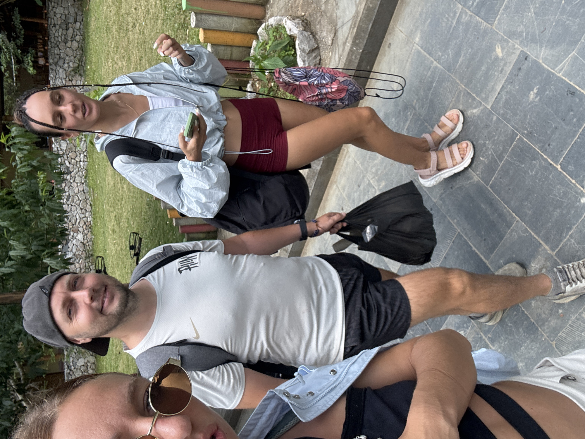 Three people stand on a paved surface, likely in Mèo Vạc Commune, Vietnam. The woman in the foreground wears sunglasses and a blue denim shirt over a black top. A man next to her is dressed in a white Nike T-shirt, black shorts, a gray cap, and sneakers, holding a black plastic bag. A second woman, standing farther back, is wearing a light blue jacket, red shorts, and pink sandals, holding a phone. Backpacks are visible on the backs of both women and the man. The background includes grass and a low wooden fence.