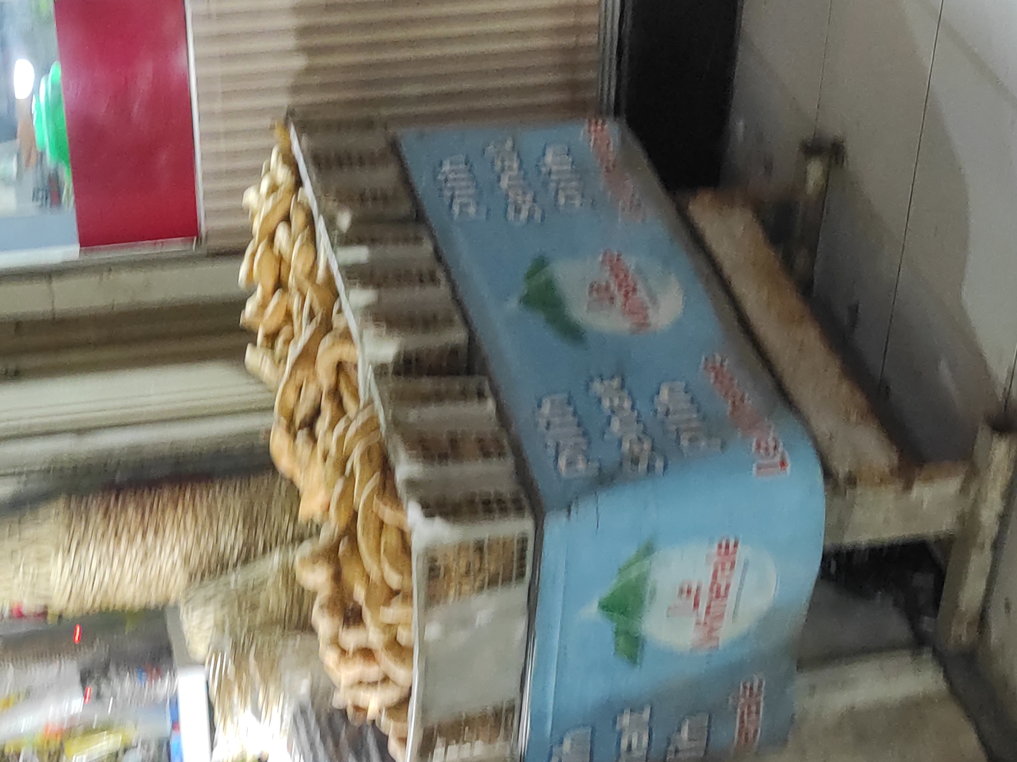 A street-side food cart is shown in an outdoor market setting. The cart is light blue with a darker blue base. The upper section of the cart holds a large quantity of golden-brown, crescent-shaped fried pastries. A white woven basket with a checkered pattern is filled with these pastries. Text on the blue section of the cart is partially visible and appears to be in Indonesian, with some words like "Pilih" (Choose) and possibly a brand name like "Le Minerale" visible on a white circular logo. The cart is positioned on a tiled surface. In the background, there are vertical striped walls, possibly part of a building or stall, and out-of-focus elements that suggest a busy market environment. The overall lighting indicates it may be evening or a dimly lit area. The image is blurry, suggesting it was taken with a handheld camera in motion or low light conditions. There are no people clearly visible in the frame.