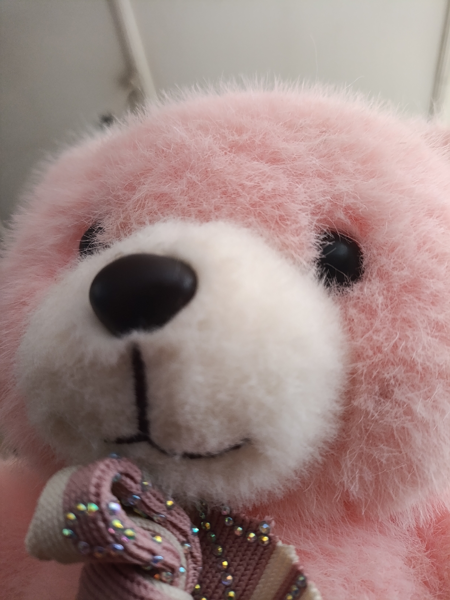 A close-up shot focuses on the face of a pink teddy bear. The bear's fur is soft and fluffy, with a lighter cream-colored muzzle. Its black button eyes are visible, and a dark, heart-shaped nose sits prominently on its snout. Below the nose, a vertical dark line suggests the bear's mouth. The bear is wearing a bow around its neck, made of striped fabric in shades of pink and cream, adorned with small, multicolored iridescent embellishments that catch the light. The background is out of focus, appearing as a soft, muted gray, with hints of vertical lines that might suggest a wall or doorway. The lighting is soft and diffused, casting no harsh shadows. The image evokes a sense of gentle affection and childlike wonder.