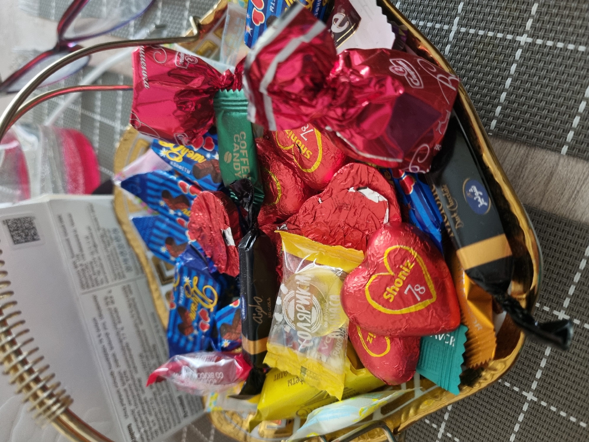 A close-up shot shows a heart-shaped gold-colored basket filled with a variety of candies. The candies are wrapped in different colors, including red, blue, yellow, green, and black. Some of the wrappers have text on them, including the words "Shoniz 78", "Coffee Candy", and "Kolyariki". There is also a box of dark chocolate. The basket is sitting on a textured surface. To the left of the basket is a spiral notebook with a white paper page that has black text on it. In the upper left corner of the image are a pair of eyeglasses. It is not possible to determine the location in the city of Sochi, Russia from the information provided.