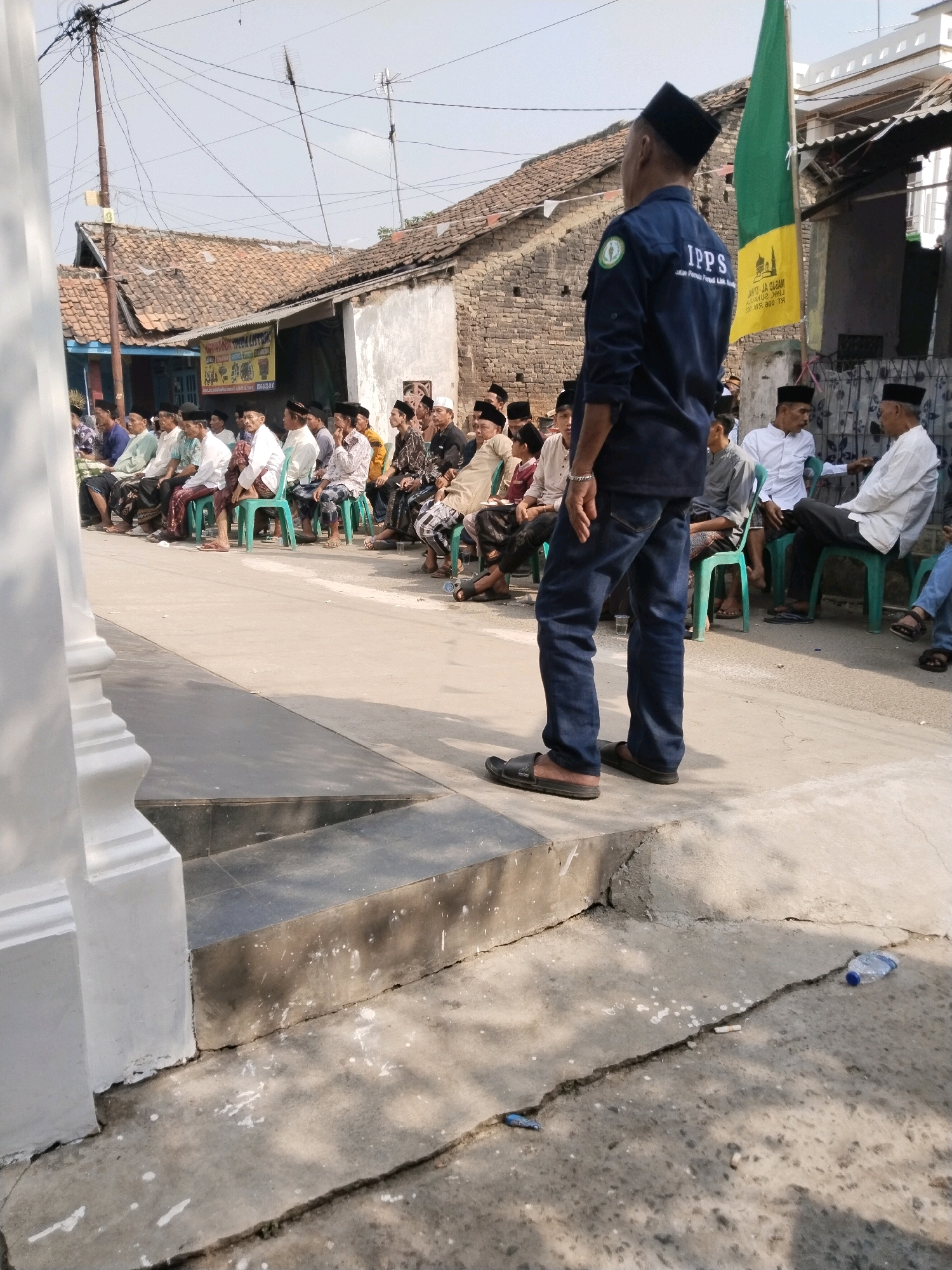 An outdoor medium shot captures a man in a dark blue uniform with "IPPS" embroidered on the back, facing away from the camera and towards a gathering of men seated on green plastic chairs. The man's uniform also features a circular green and white logo. He is wearing dark blue jeans and dark sandals.
The seated men, all wearing traditional Indonesian attire including peci hats, are arranged in rows in front of a low-slung building with a terracotta tile roof. Many are looking towards the man in uniform, suggesting he might be addressing them or observing the gathering. A green and yellow flag with Arabic script and an image of a mosque is prominently displayed behind the seated men.
The environment appears to be a village setting in Serang, Indonesia, indicated by the style of the buildings and the attire of the people. The ground is paved with concrete, showing signs of wear and tear, including cracks and scattered debris. A white pillar is visible in the foreground on the left side of the image. The overall scene suggests an organized outdoor event, possibly a religious or community meeting, taking place under a bright, clear sky.