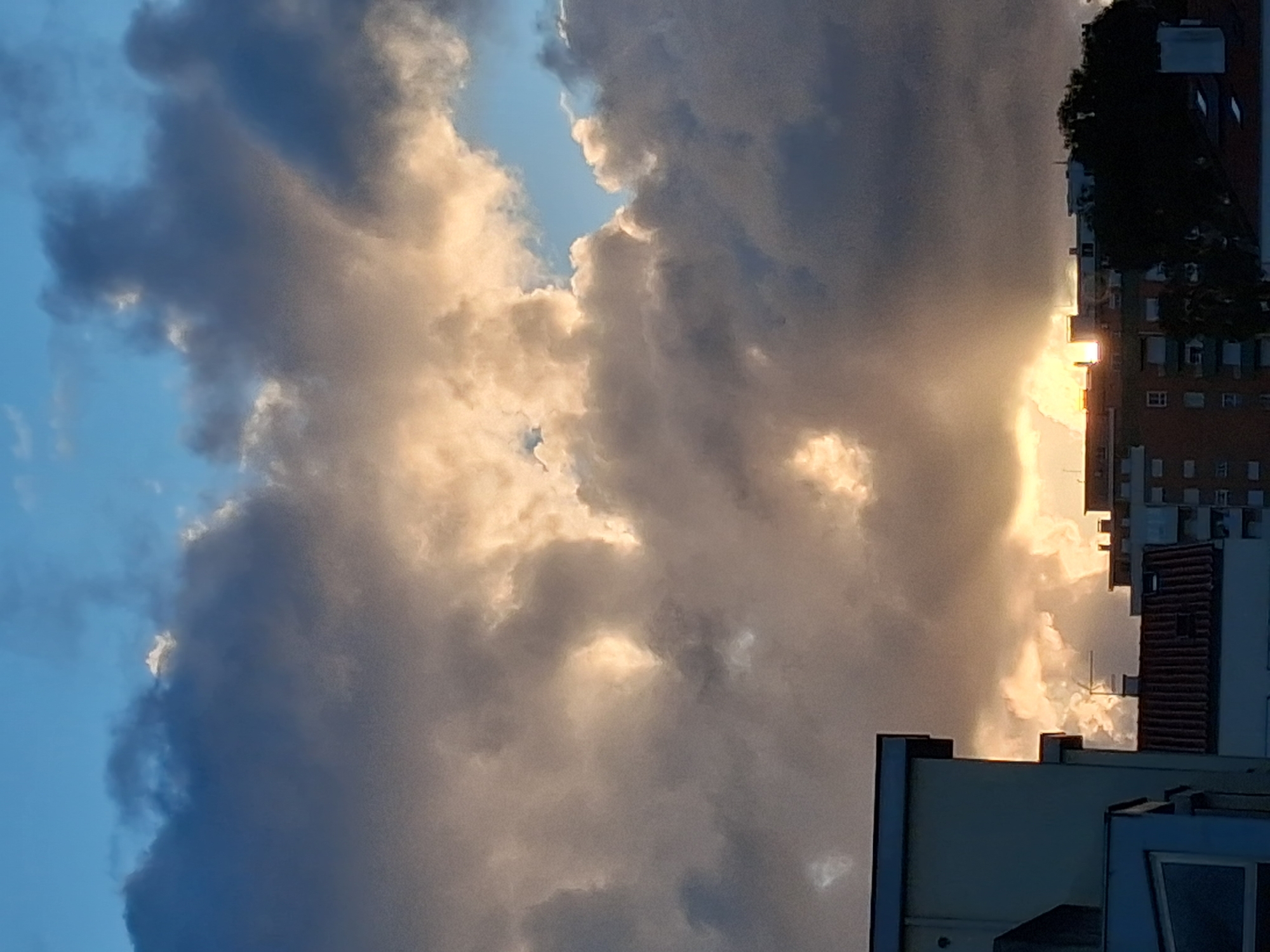 The image shows a section of the sky filled with dark, billowy clouds with light shining through gaps in the clouds. Buildings are visible on the right side of the image, one with a dark facade. The image may be taken from Figueira da Foz, Portugal, but it is hard to determine. There are no people, subjects or actions in the image.