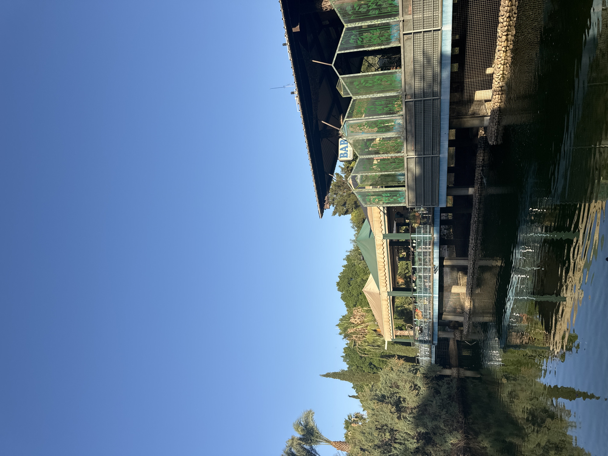 The image depicts an outdoor dining establishment built over a body of water, likely a lake or pond, with lush greenery and trees surrounding it. The architecture features covered seating areas with tan and green awnings, supported by pillars. A prominent blue railing encloses an elevated walkway or patio area in front of the building, which has decorative glass panels with a bamboo motif. A sign with the word "BAR" is visible above the entrance. The water's surface reflects the structures and the surrounding vegetation, creating a serene and somewhat tropical ambiance. The sky is a clear, bright blue.
While the image showcases a scenic location with a restaurant and water feature, it does not contain any specific visual cues or landmarks that would definitively place it in the city of Murcia, Spain. Without additional context or identifying features unique to Murcia, it is impossible to confirm its location within that city.