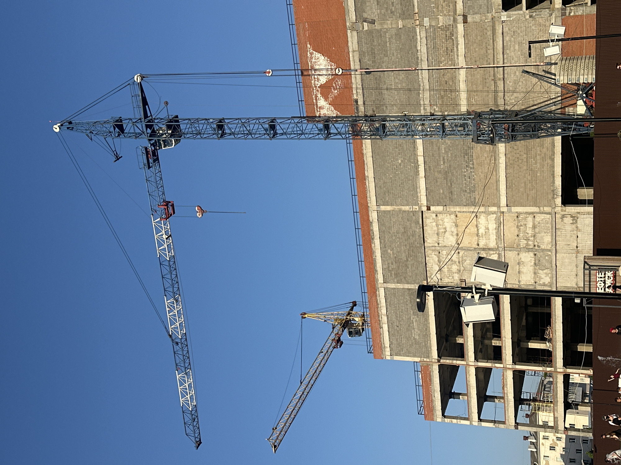 The image shows a construction site in Neftekamsk, Russia. Two cranes are visible against a clear blue sky. One crane is taller and has a blue-grey color, while the other is shorter and yellow. Both cranes are positioned next to a tall, unfinished building with a concrete structure and some brick sections. At the base of the building, some people are present, standing in front of a dark brown wall with a sign visible that spells "COFE KICK". There are other buildings in the background beyond the construction site.