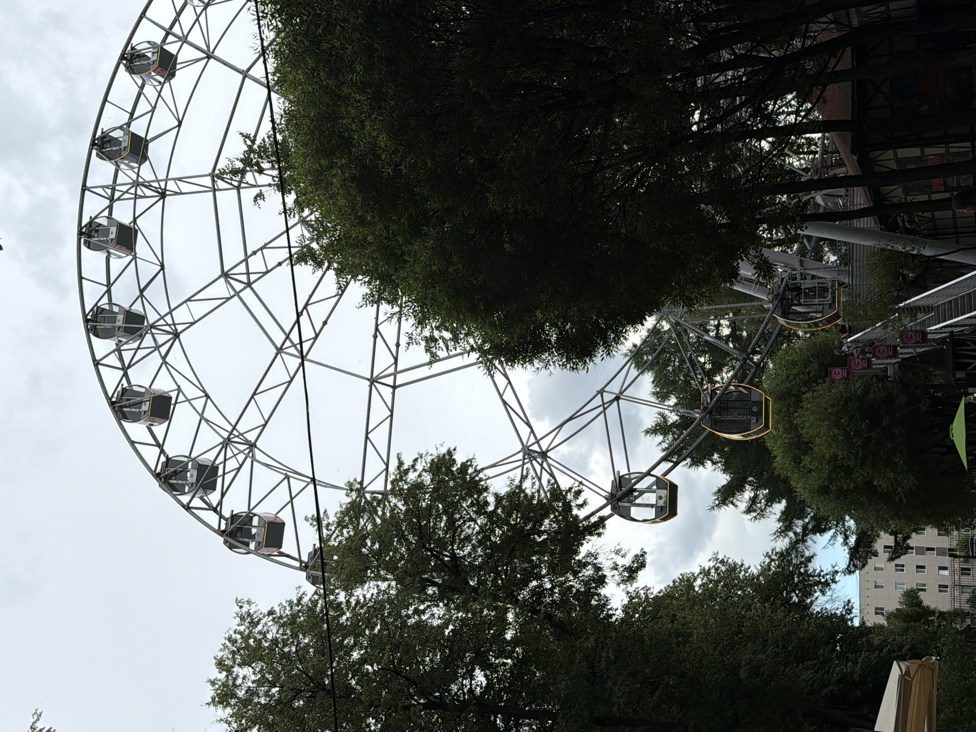 The image captures a Ferris wheel at an amusement park, likely located in the city of Perm, Russia. The Ferris wheel is partially obscured by lush green trees. The sky is overcast with a pale, cloudy atmosphere. Several gondolas are visible on the wheel, with some appearing dark gray and others with yellow trim. The structure of the Ferris wheel is intricate, composed of a network of metal beams and supports. In the background, a light-colored apartment building with multiple windows can be seen, suggesting an urban setting. To the right of the Ferris wheel, there are smaller amusement park structures with brown roofs and visible wooden elements. A metal staircase with banners leads upwards toward the Ferris wheel. In the foreground, parts of trees and some park amenities are visible, including a green umbrella. The overall impression is a daytime scene at an amusement park, likely during a period of cloudy weather.