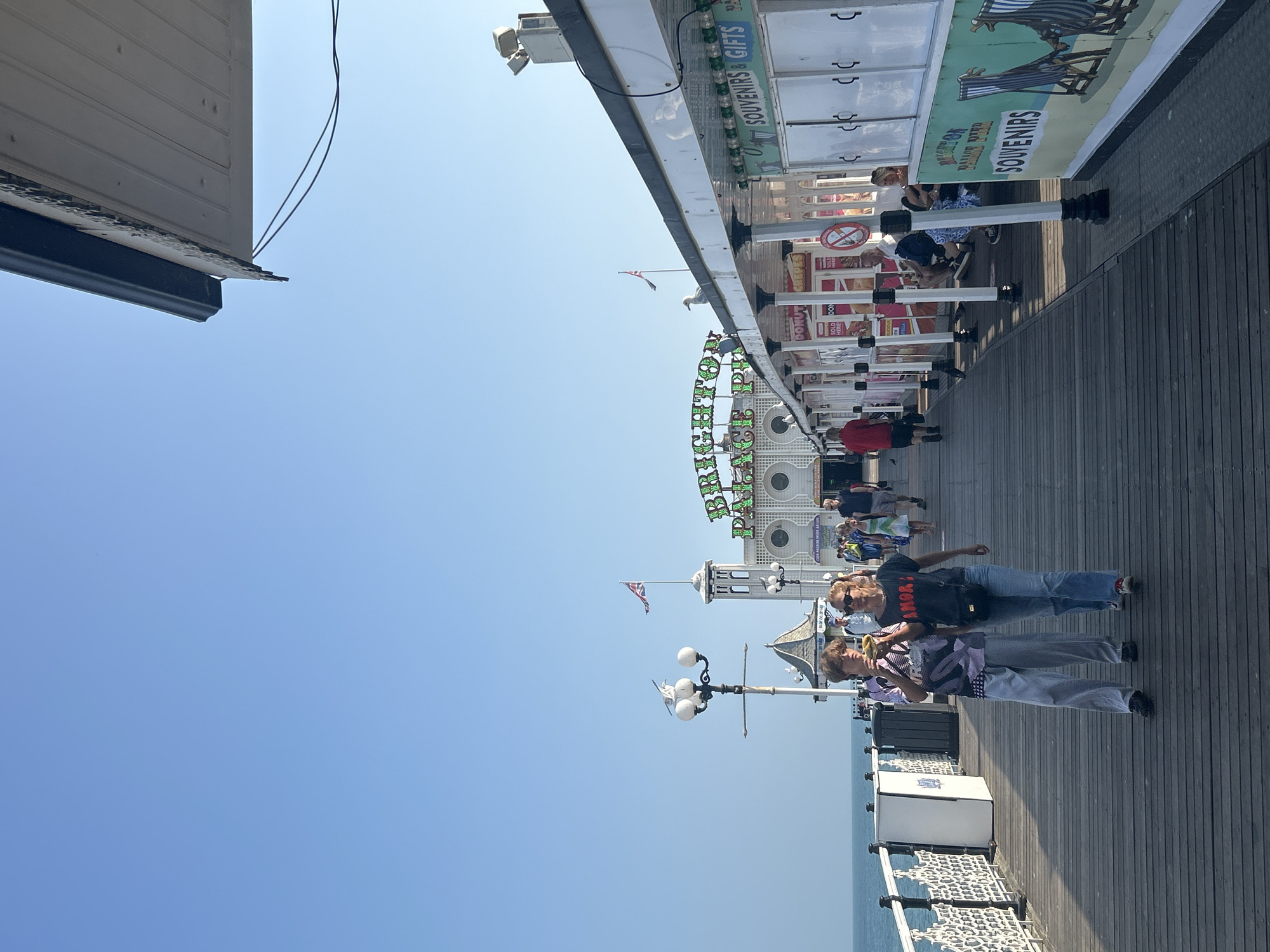 The image depicts Brighton Palace Pier in Brighton, United Kingdom, on a clear, sunny day. The pier extends out over the sea, with a wooden boardwalk lined with various stalls and attractions. In the foreground, two individuals are walking towards the camera. The person on the left, appearing younger, is wearing a purple and white patterned shirt and light grey trousers, and is holding a yellow object to their mouth. The person on the right, appearing older, is wearing a dark t-shirt with the word "AMOR" in red lettering and blue jeans.
Further down the pier, other people are visible, some seated at stalls and others walking. The stalls offer a variety of goods, including donuts, crepes, and souvenirs, as indicated by signage. A large green sign with illuminated text spelling "BRIGHTON PALACE PIER" is prominently displayed in the background. A Union Jack flag flies from a tower structure. The sea is visible to the left of the pier, and the sky is a clear, bright blue. The architecture of the pier includes white pillars supporting covered walkways and stalls with white paneling and green trim.
