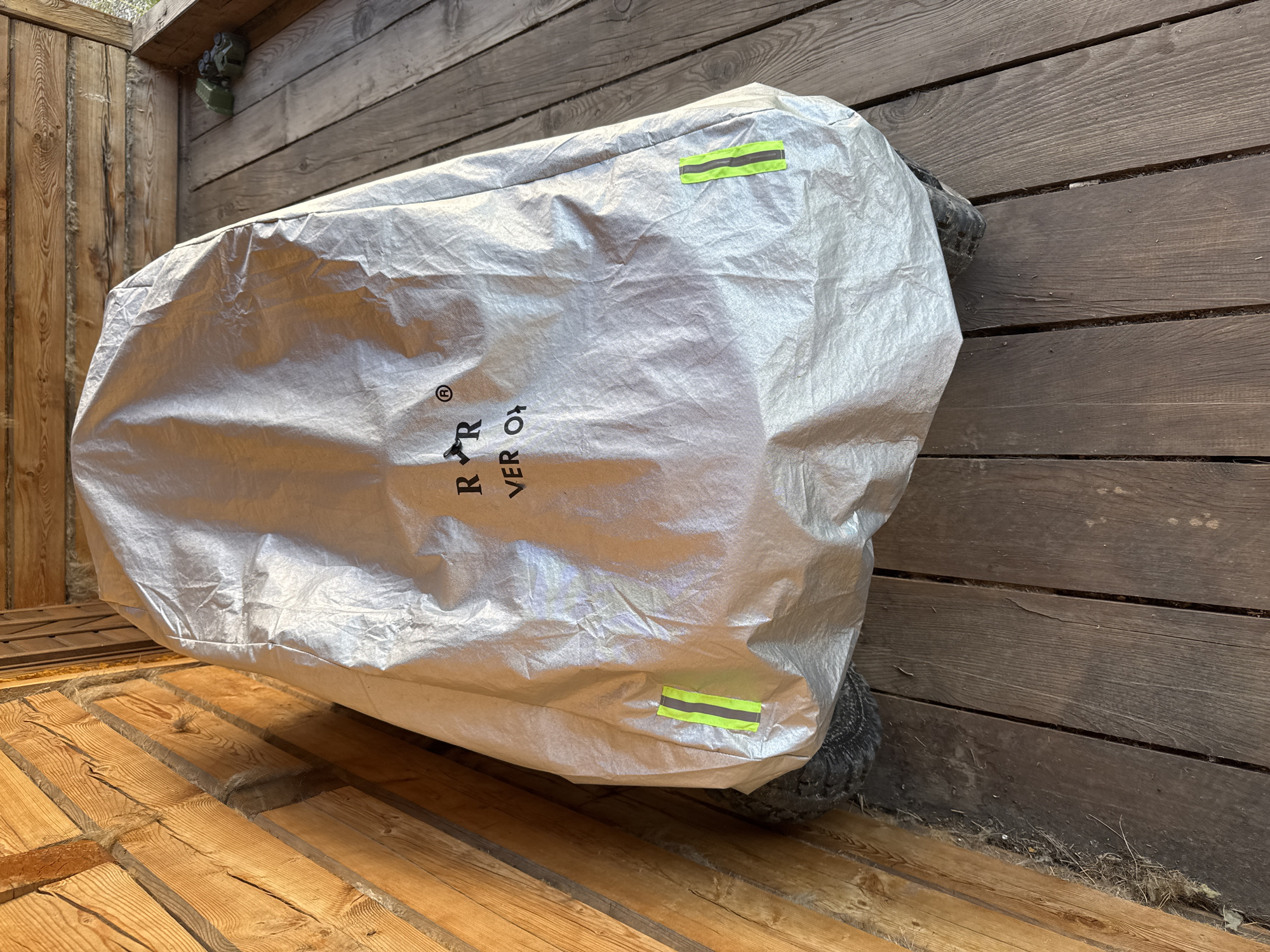 A silver-colored cover is draped over an object, likely a vehicle given the visible tires. The cover features black lettering and a logo, appearing to read "R R VER O". Two bright yellow reflective strips are attached to the sides of the cover. The object is situated on a wooden deck, next to a wooden structure with visible logs and beams. The environment appears to be outdoors, possibly in a covered area like a porch or shed. There are no people visible in the image.
