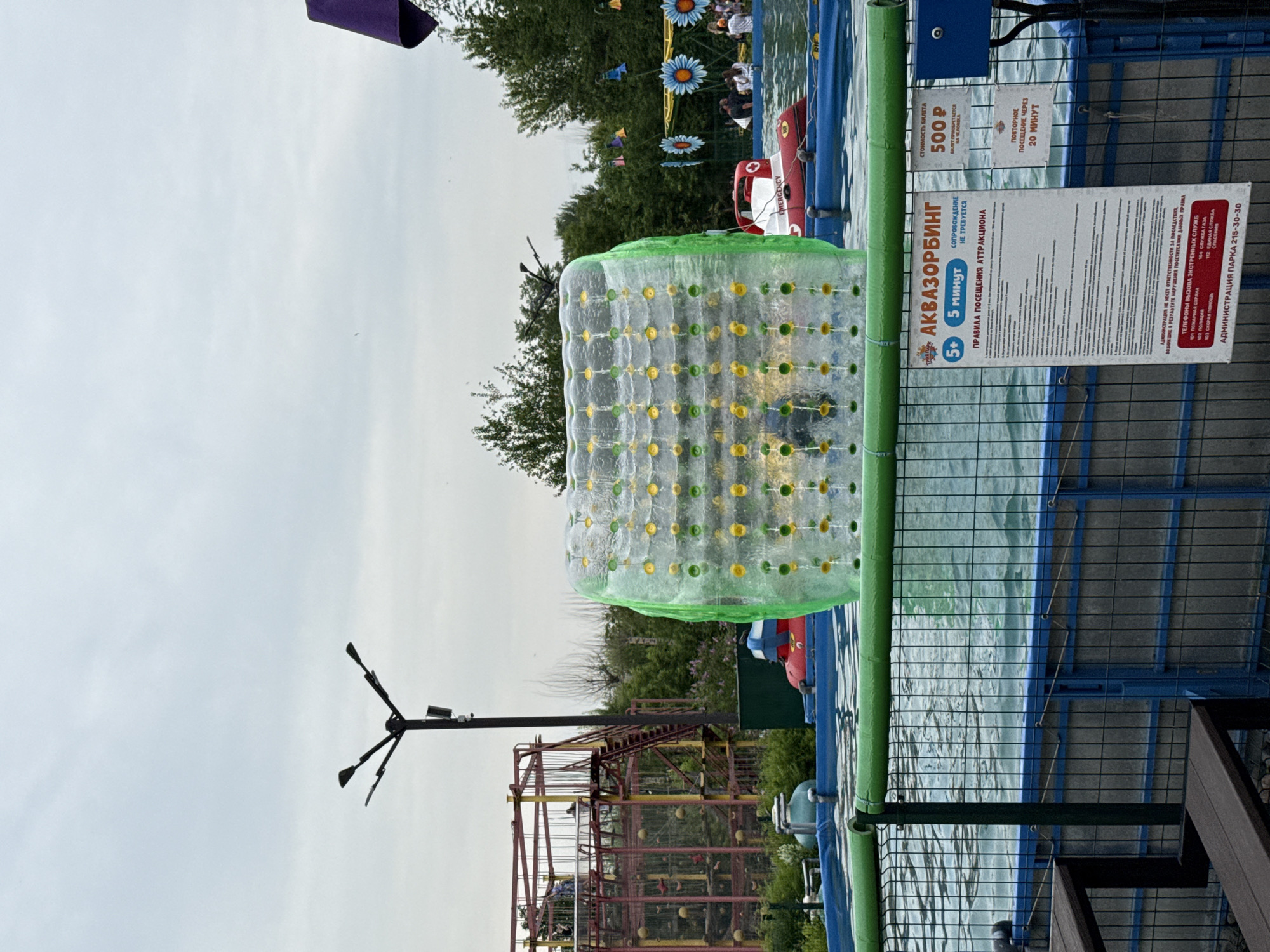The image captures an outdoor amusement park scene on a cloudy day. In the foreground, a large, transparent inflatable cylinder, likely an "aquazorbing" attraction, floats in a blue pool. The cylinder is green around the edges and filled with yellow and green dots. A sign is attached to a fence in front of the pool, providing information about the attraction. The sign is written in Russian and mentions "AQUAZORBING," "5 MINUTES," and the cost of a ticket. In the background, a red and white inflatable boat with the word "EMERGENCY" printed on its side is partially visible in the pool. To the left, a tall, multi-level structure with red metal framing and stairs suggests another attraction, possibly a climbing wall or ropes course, with people visible at higher levels. Several trees and other park features, including decorative flowers and colorful hanging objects, are scattered throughout the background, suggesting a vibrant park environment. The overall impression is a recreational setting, likely a water park or amusement park. Given the presence of Russian text on the sign and the general aesthetic, this scene is consistent with a location in Krasnoyarsk, Russia.