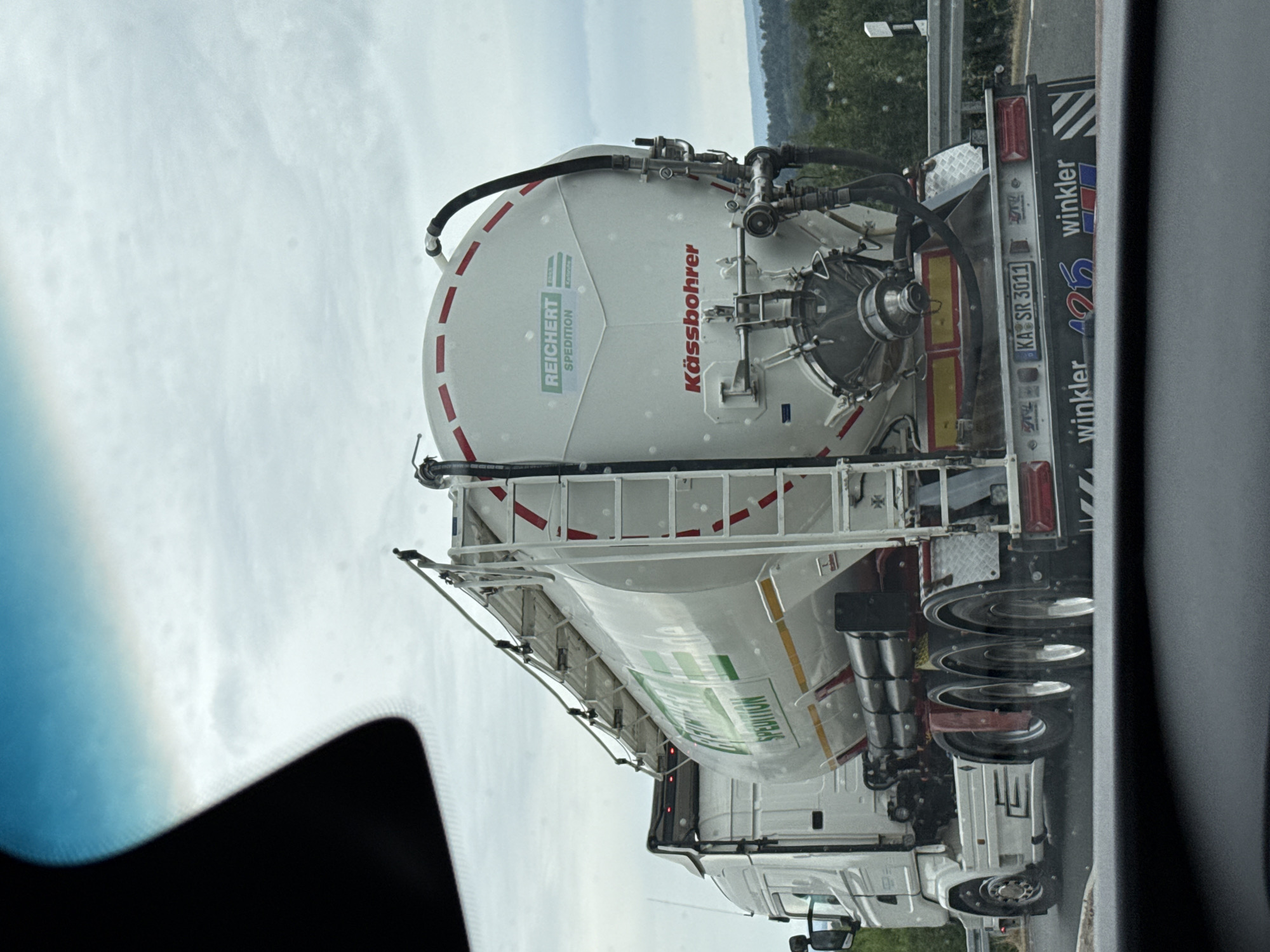 The image shows the rear of a white tanker truck driving on a road. The truck has a large cylindrical tank with "REICHERT SPEDITION" and "Kässbohrer" logos. It has a ladder on the side and various hoses and valves. The license plate reads "KA SR 3011". In the background, there are trees and a cloudy sky. The perspective suggests the photo was taken from inside another vehicle. The location is identified as Bad Sachsa, Germany.
