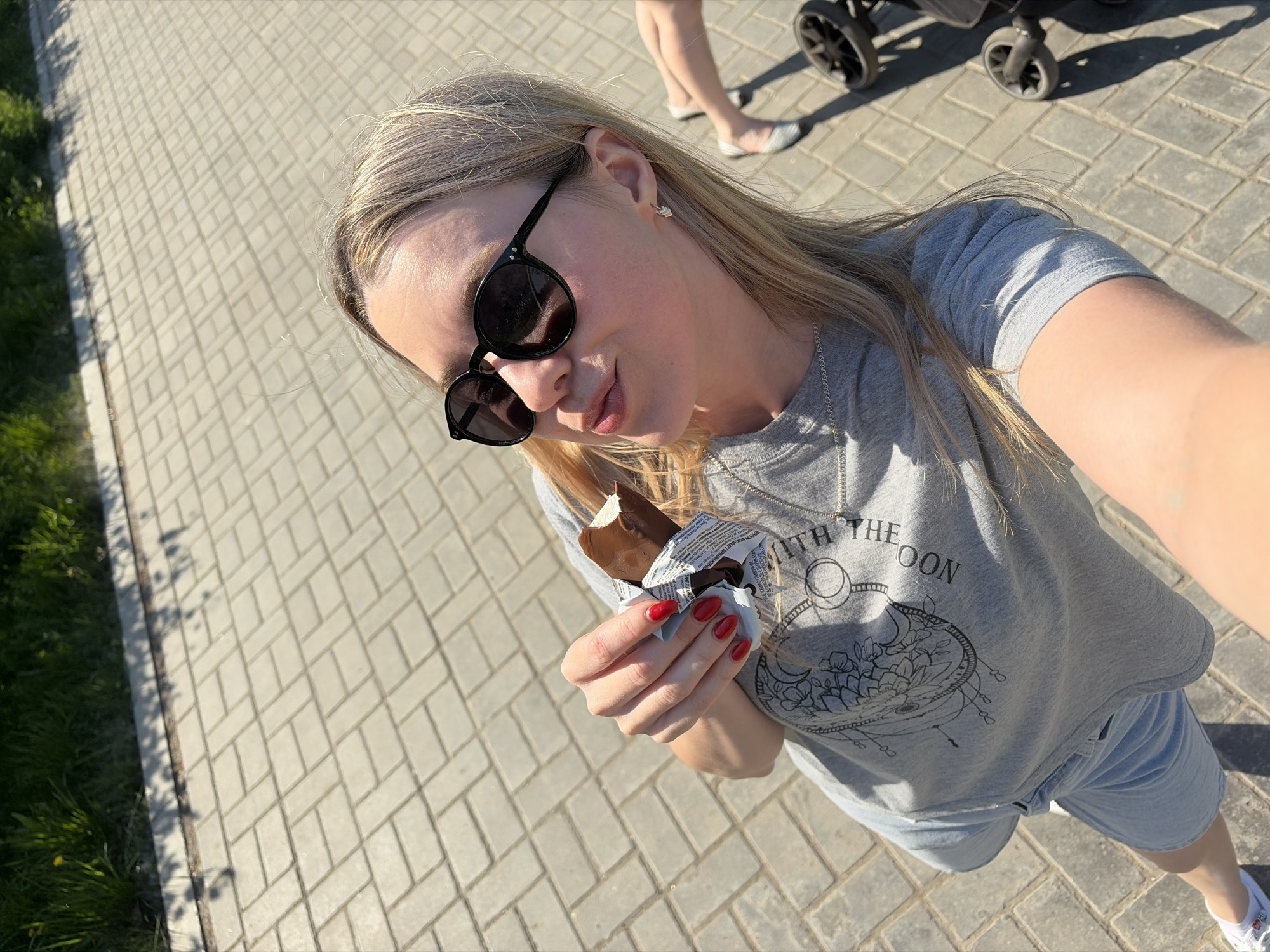 A high-angle, full shot depicts a young woman eating an ice cream bar while outdoors. She is standing on a paved walkway, clad in a grey t-shirt with a circular design, light blue shorts, and white sneakers. She is wearing black sunglasses and a silver necklace. Her nails are painted a bright red. She holds the half-eaten ice cream bar in her left hand.
In the background, a portion of a baby stroller is visible, with a person's legs nearby. The environment consists of a paved walkway with grass on the left. The location is Chaikovsky, Russia.