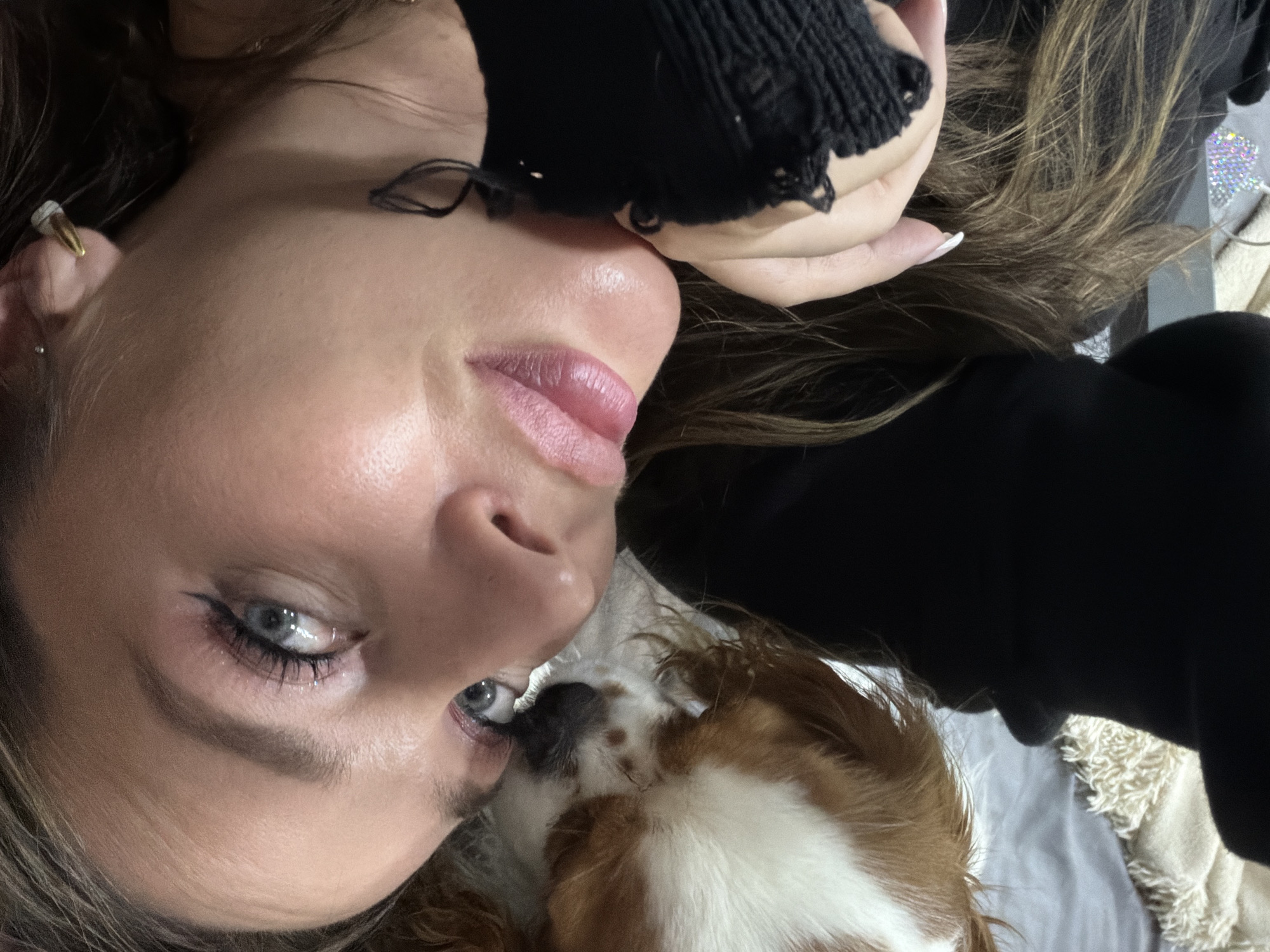 A close-up, top-down shot captures a woman and a small dog resting together indoors in Chaikovsky, Russia. The woman, with light skin, blue eyes, and dark brown hair, is positioned on her back, looking upwards towards the camera. She has eyeliner and pink-tinted lips, and a gold earring with a white tip in her right ear, along with a small silver stud. She is wearing a black long-sleeved top, possibly a sweater, with distressed detailing on the sleeve.
A small dog, appearing to be a Cavalier King Charles Spaniel with white and brown fur, rests its head closely against the woman's chin, also looking upwards. The background suggests a comfortable indoor setting, possibly a bed or couch, with light-colored, textured blankets or fabrics. A sparkling, decorative object is partially visible in the upper right corner. The overall scene conveys a feeling of intimacy, companionship, and peace between the woman and her pet. No visible text is present in the image.