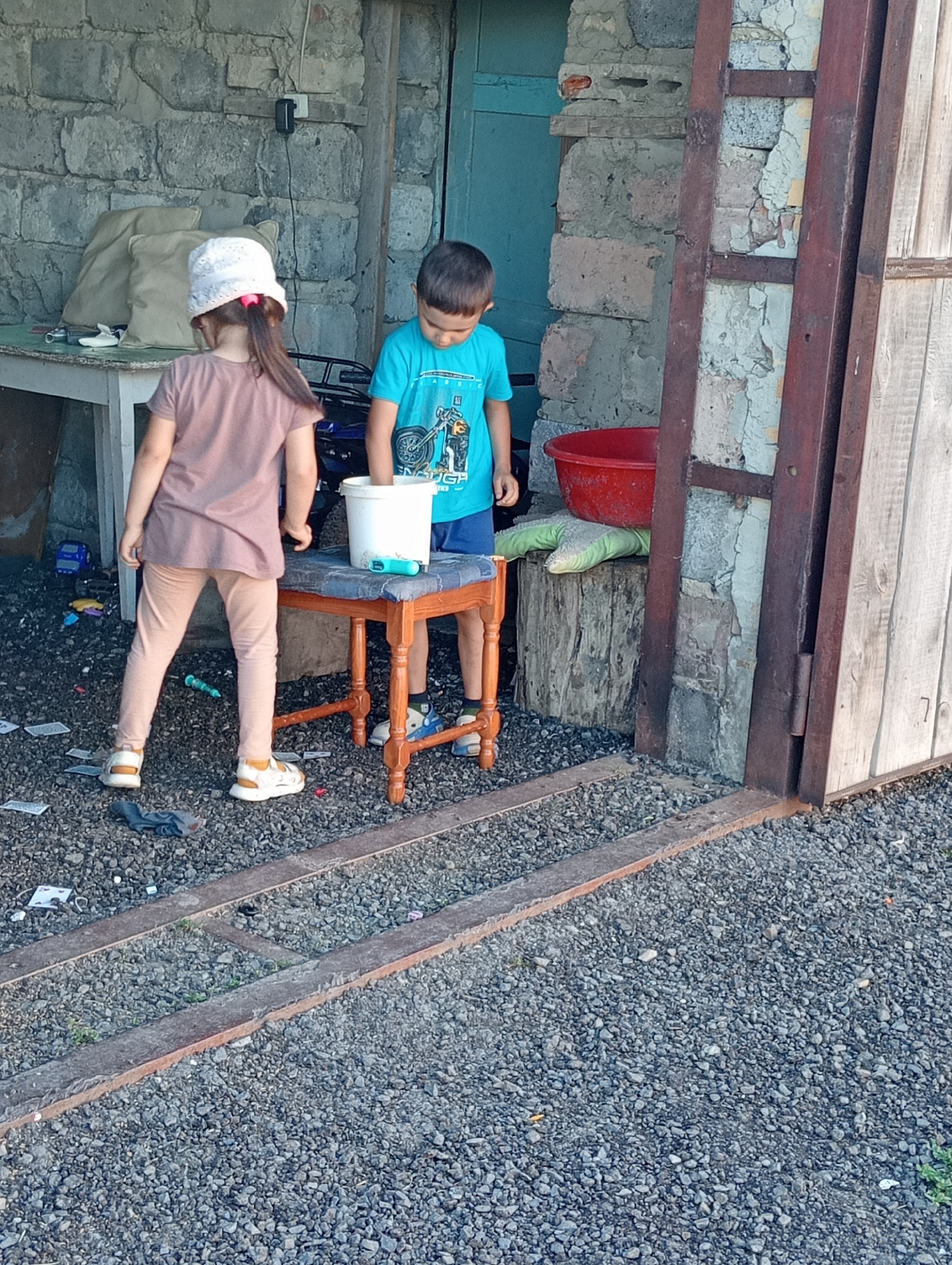 In an outdoor setting with a gravel ground, two children are present. A young girl wearing a pink shirt, beige leggings, and a white hat stands on the left, facing away from the camera. A young boy in a blue t-shirt and dark shorts stands to her right, also facing away. The boy is looking down at a white bucket placed on a small wooden stool. Behind the children, a stone wall with a doorway is visible. To the right of the doorway, a large wooden door is present. The scene appears to be in a rural or industrial area, possibly a backyard or an abandoned structure. There are various objects scattered on the ground, including toys and debris. The lighting suggests it is daytime.