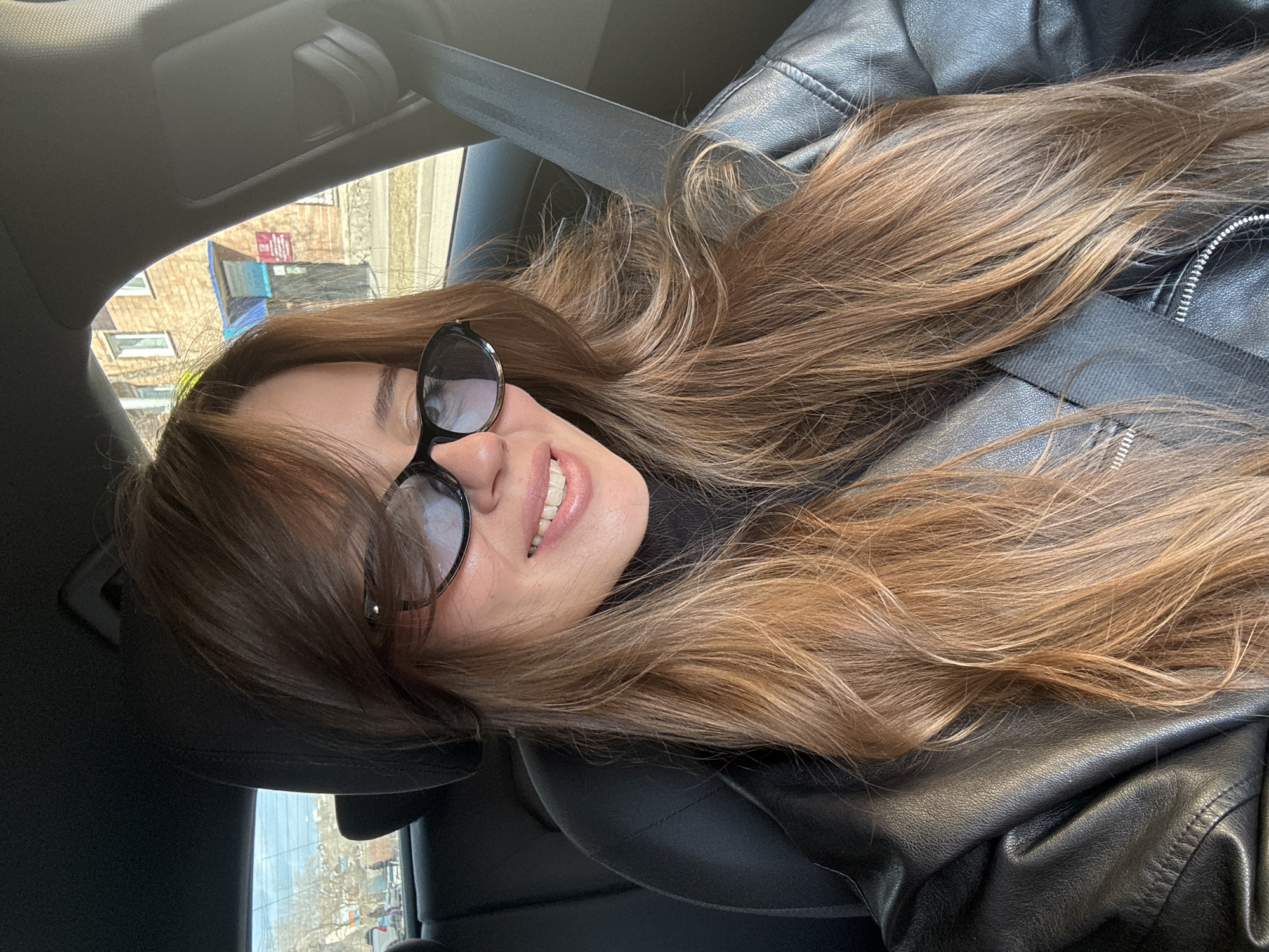 A person wearing sunglasses and a black leather jacket is photographed inside a car. The person is wearing a seatbelt. Outside the car window, there is a building with a blue roof. This building is located in Krasnoyarsk, Russia. The image is taken at an angle. The person has long brown hair.