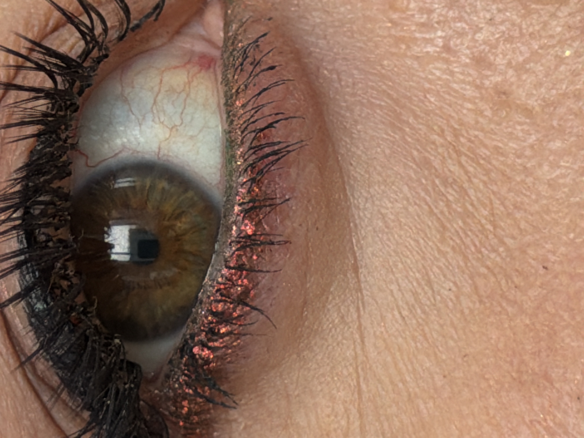 A close-up of a human eye with brown iris, surrounded by heavily mascaraed eyelashes. The eyelid shows skin texture and makeup with reddish-gold glitter. There is no indication that this photo was taken in Krasnoyarsk, Russia.