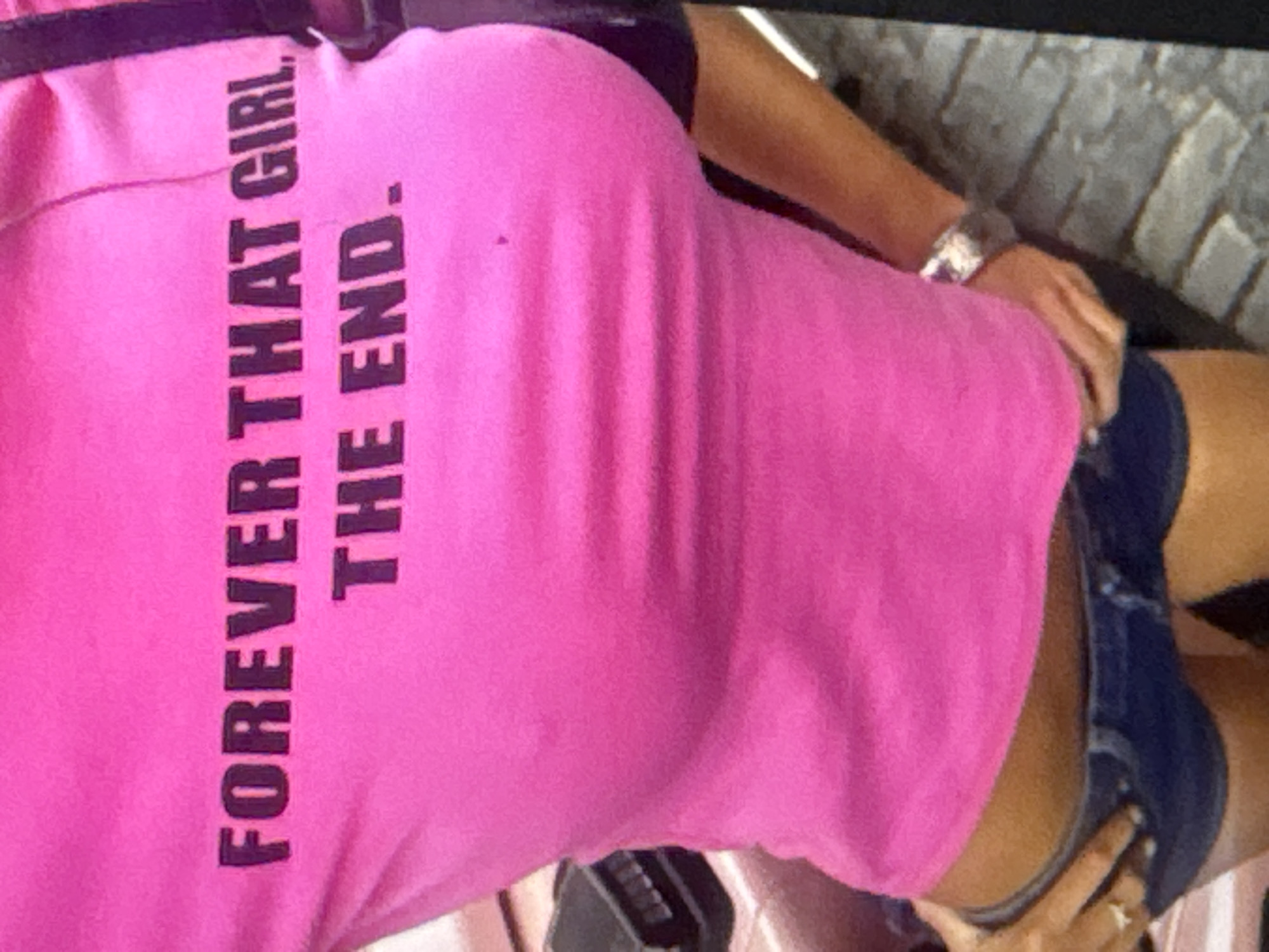 The media file shows a close-up of a person wearing a bright pink t-shirt with dark text printed vertically. The text reads "FOREVER THAT GIRL. THE END.". The t-shirt appears to be made of a ribbed fabric. The person is also wearing dark blue denim shorts. A silver watch is visible on the person's left wrist. The background is blurred but appears to be an outdoor setting with textured ground, possibly cobblestone or brick, and hints of a wall. The lighting suggests it is daytime. The image is taken from the side and slightly from behind, focusing on the t-shirt and the person's torso and upper legs.