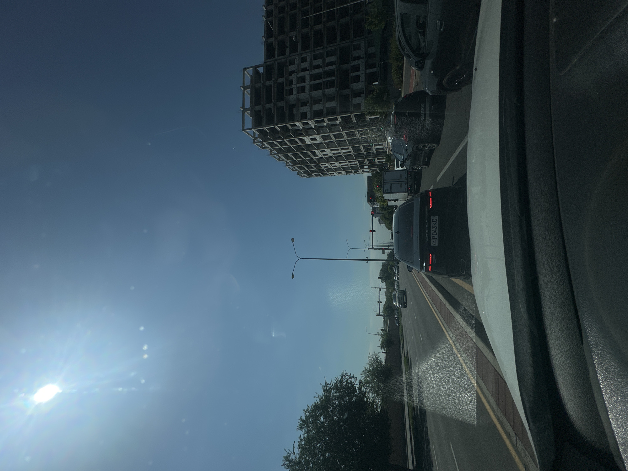 This is a low-angle, roadside shot taken from the interior of a vehicle, looking forward. The scene captures a traffic jam on a multi-lane road, likely a major thoroughfare in Tashkent, Uzbekistan, given the license plate format visible on the black Chevrolet sedan in the foreground. The sky is a clear, bright blue with the sun positioned in the upper left quadrant, creating lens flare and bright streaks across the windshield.
In the immediate foreground, the dashboard and windshield of the vehicle from which the photo is taken are visible. Ahead, several cars are stalled in traffic, including the black Chevrolet with a license plate displaying "01 PD043KC". Beyond that, other vehicles, including a white truck and several SUVs, are also present in the traffic.
To the right of the road, an unfinished, multi-story concrete building with exposed rebar and open window frames dominates the middle ground. This skeletal structure suggests ongoing construction. Streetlights line the road, and traffic signals are visible further down.
On the left side of the road, a large, mature tree with dark green foliage provides shade, and a brick wall runs along the edge of the road. The road itself is asphalt with clear lane markings. The environment appears to be an urban or suburban setting, characterized by a mix of infrastructure, residential or commercial development, and greenery. The overall impression is a typical city scene on a sunny day, with the added visual interest of the stark, unfinished building against the bright sky.