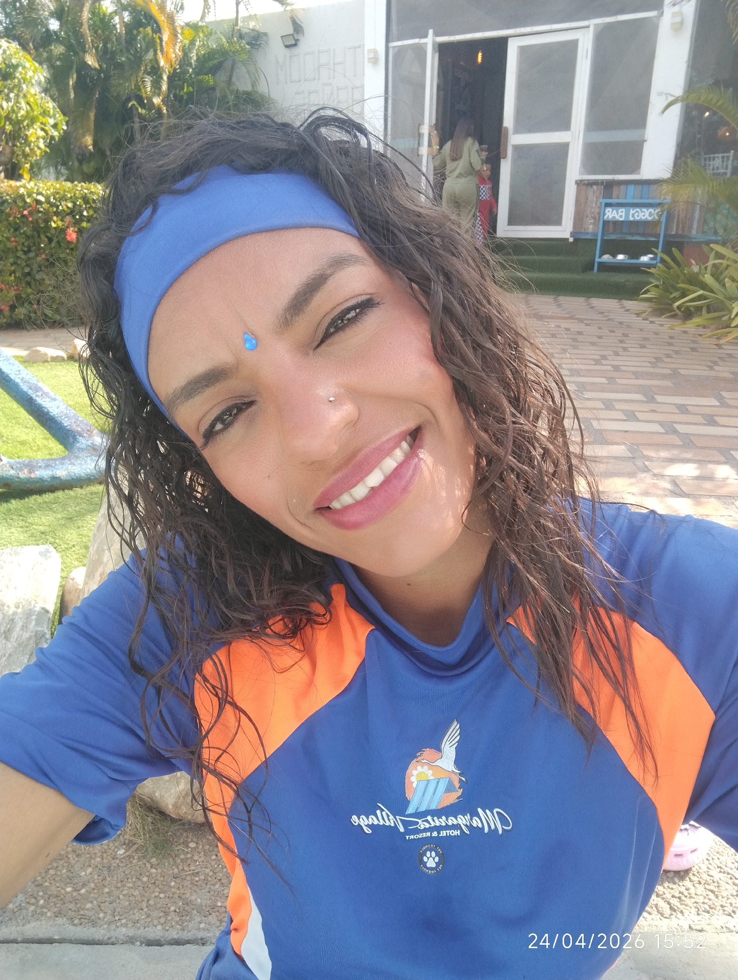 A smiling woman with dark, curly hair and a blue headband takes a selfie outdoors. She wears a blue and orange long-sleeved shirt featuring a white logo with a bird, a paw print, and mirrored text that appears to read "MOZART HOTEL & RESORT". A blue teardrop-shaped adornment is visible on her forehead, and she has a small silver nose piercing.
In the background, a white building with large glass doors is visible. Text on the building itself suggests "MOZART HOTEL & RESORT". A blue, wooden bench-like structure with "MOZART BAR" written in mirrored text ("РАВ ЯЗООМ") is positioned to the right of the doors. Two other individuals, an adult in a green outfit and a smaller person, are partially visible near the doorway, entering or exiting the building.
The setting includes a vibrant outdoor area with green artificial turf, paved pathways, rocks, and lush greenery including palm trees. A blue, anchor-like decorative element is partially visible on the left. The scene is brightly lit by sunlight, indicating fair weather. A timestamp "24/04/2026 15:52" is present at the bottom right, suggesting the photo was taken in the late afternoon. The overall atmosphere appears cheerful and relaxed, typical of a resort or leisure establishment.