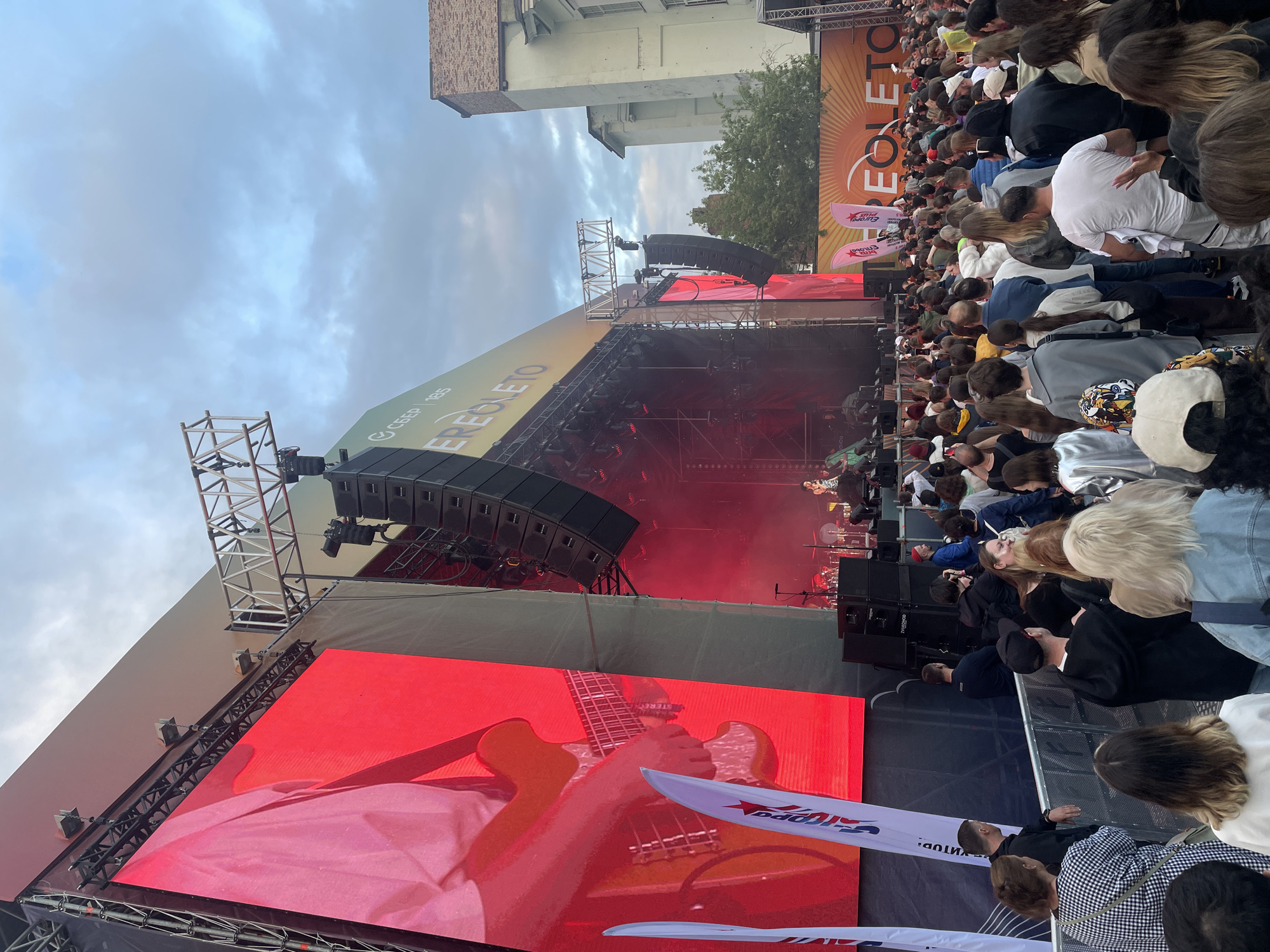 This image captures a daytime outdoor concert event, likely in Saint Petersburg, Russia, given the visible Cyrillic text "EREGLETO" and sponsorship logos including "СБЕР 185". A large stage is set up with prominent LED screens displaying a close-up of a person playing an electric guitar. The stage is equipped with numerous speakers and lighting rigs. A crowd of spectators, diverse in age and gender, fills the area in front of the stage, many holding up phones to capture the performance. The environment appears to be an urban setting, with buildings visible in the background to the right. The sky is overcast with clouds. Various promotional flags and banners are placed around the venue.