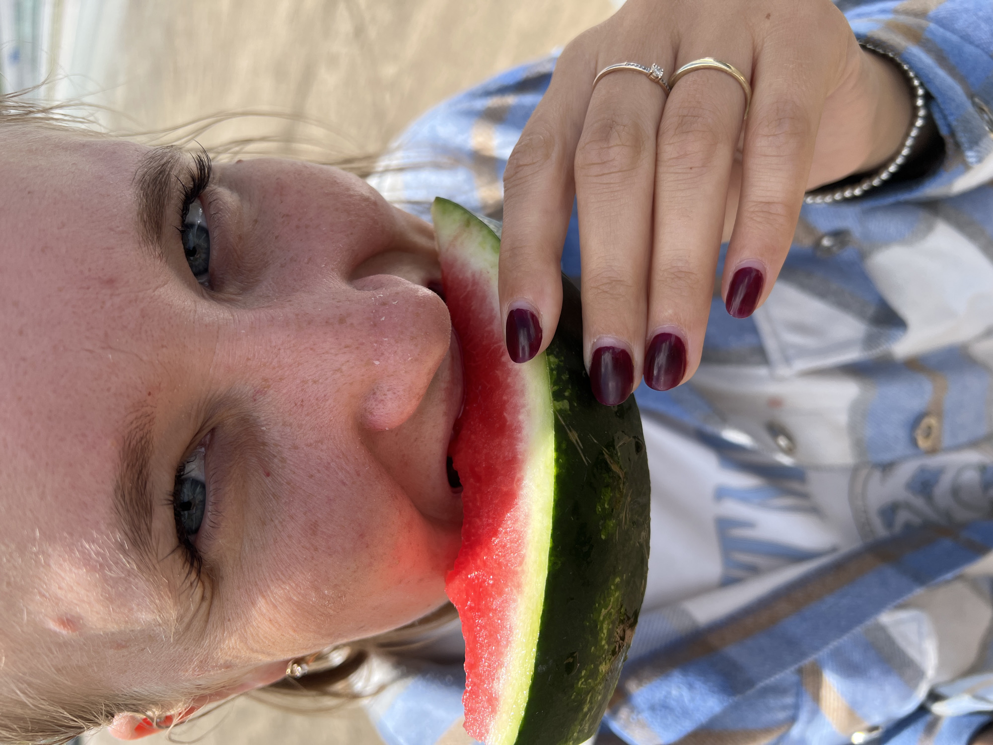 In this close-up, vertically oriented image, a person is shown eating a slice of watermelon. The individual's face is partially visible from a low angle, with their eyes looking upwards and to the left. They have fair skin with freckles and blue eyes. Their mouth is open, taking a bite of the watermelon, which has a bright red interior and a green rind.
The person is wearing a blue and white plaid shirt. Their right hand is visible, holding the watermelon slice. The hand has dark red nail polish, and there are two rings on the ring finger, one appearing to be a simple band and the other a thin band with a small gem.
The background is blurred, but appears to be an outdoor setting, possibly a beach or picnic area, with light-colored sand or fabric. The overall lighting suggests it is daytime, likely sunny, given the brightness. The scene evokes a sense of casual enjoyment and refreshment on a warm day. No text is visible in the image. The location is identified as Alexandrovka, Russia.