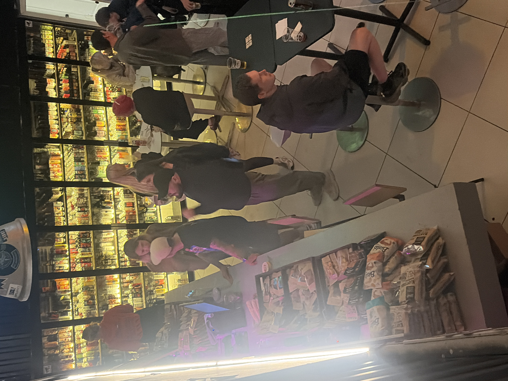 The image depicts an indoor scene, possibly a bar or a retail establishment with a refrigerated display of beverages. Several individuals are present, some interacting with each other and others standing or sitting. In the foreground, a counter is visible with a variety of packaged goods, suggesting it might be a service area. The lighting is dim with prominent neon accents. The overall atmosphere appears casual. The location of Saint Petersburg, Russia is not evident from the visual information in the image.