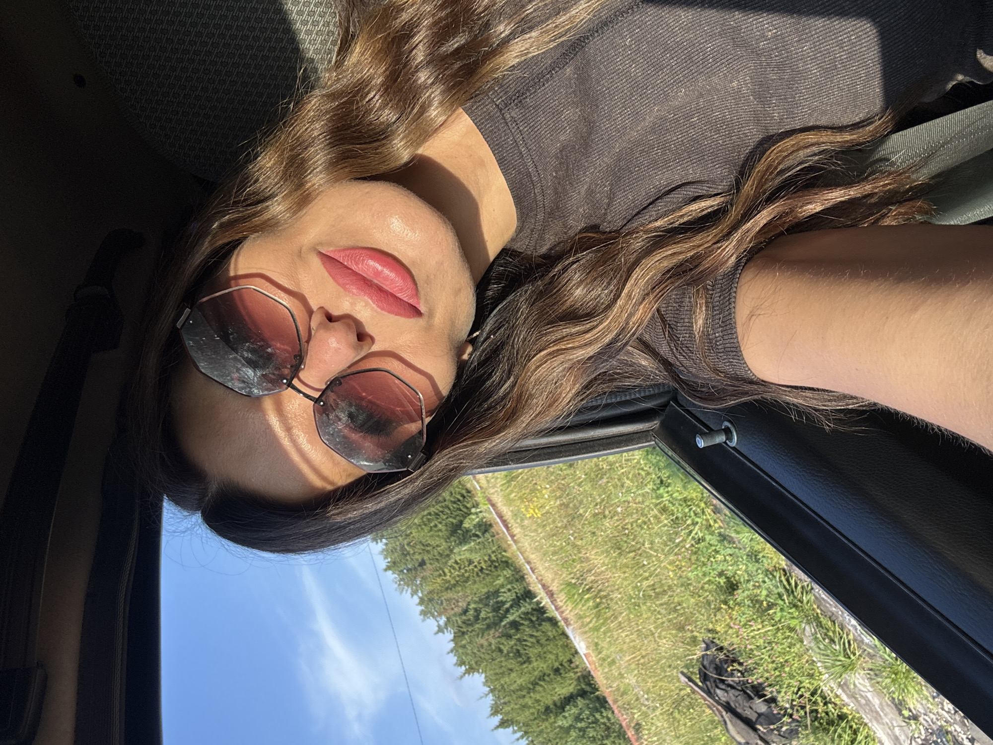 A woman with long, wavy brown hair and sunglasses is taking a selfie in a car. She is wearing a dark gray t-shirt. The sun is shining, casting shadows on her face. Outside the car window, there is a grassy field with trees in the background, suggesting a rural or suburban setting. The car interior is visible, including the door panel and a part of the seat. The image does not contain any specific indicators of the city of Olkhovka, Russia.