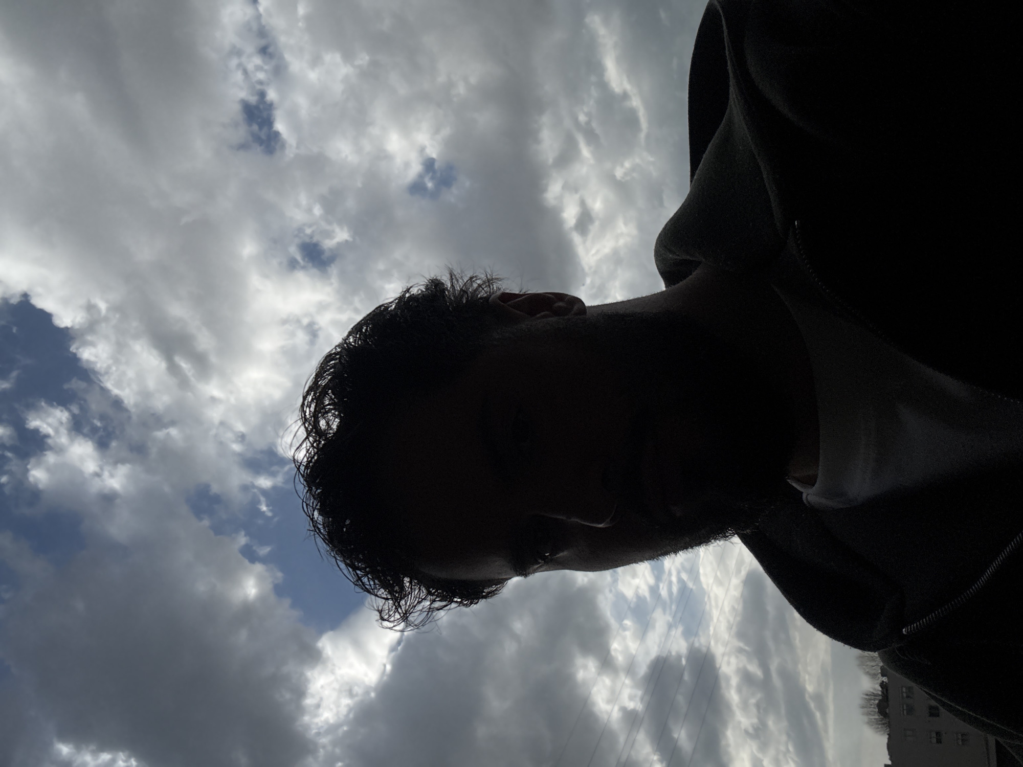 A male individual is depicted in a high-contrast, low-angle, back-lit close-up shot against a cloudy sky in Vila Nova de Gaia, Portugal. The subject's face is heavily shadowed, forming a silhouette, revealing the outline of his head, dark curly hair, a prominent nose, and the right ear. He wears a dark, possibly black or dark grey, hooded jacket with a visible zipper, over a lighter-colored, likely white, shirt. The sky behind him shows a scattered cloud formation with significant patches of blue sky. In the mid-ground on the left, multiple parallel utility wires are visible. Below these wires, indistinct outlines of urban buildings and bare trees are partially visible in the lower left quadrant. No specific actions or interactions are depicted beyond the subject's static pose, possibly a selfie.
