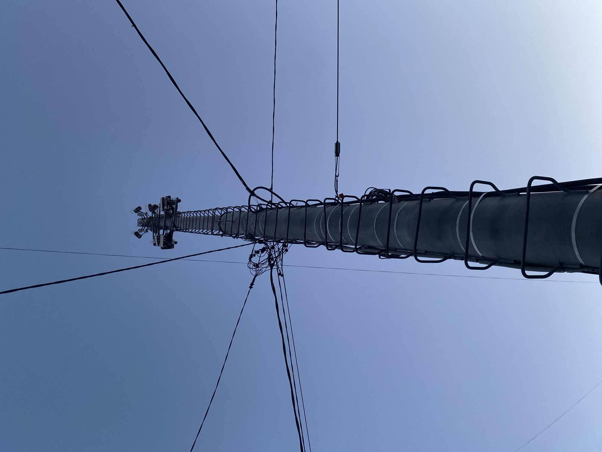 The image depicts a tall communications tower against a clear blue sky. The tower is cylindrical with ladder-like rungs running along one side. Various wires and cables are connected to the tower and extend outwards. At the top of the tower, there are multiple antennas and electronic equipment. The location is Izhevsk, Russia.
