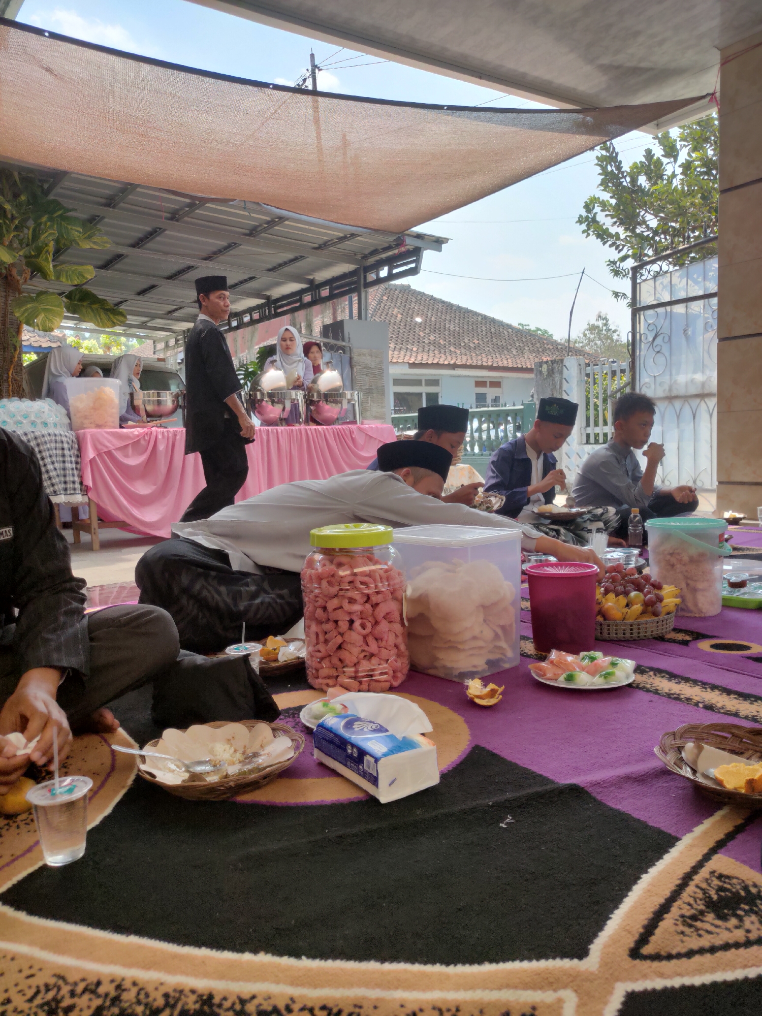 The image depicts an outdoor gathering, likely a communal meal, set in Sumedang, Indonesia. The scene is characterized by a festive, informal atmosphere. People, mostly men and some women dressed in modest attire including headscarves, are seated on a colorful patterned rug on the ground. They are engaged in eating, with plates of food, drinks, and snacks laid out before them. Several large containers of snacks, such as crackers and puffed snacks, are prominent. In the background, a buffet-style setup with chafing dishes on a pink tablecloth suggests a catered event. The environment includes a covered outdoor area with a corrugated metal roof, shading the gathering from the sun. Trees and houses with traditional tiled roofs are visible in the distance, indicating a residential or community space within Sumedang. The lighting suggests it is daytime, with natural sunlight filtering through.
