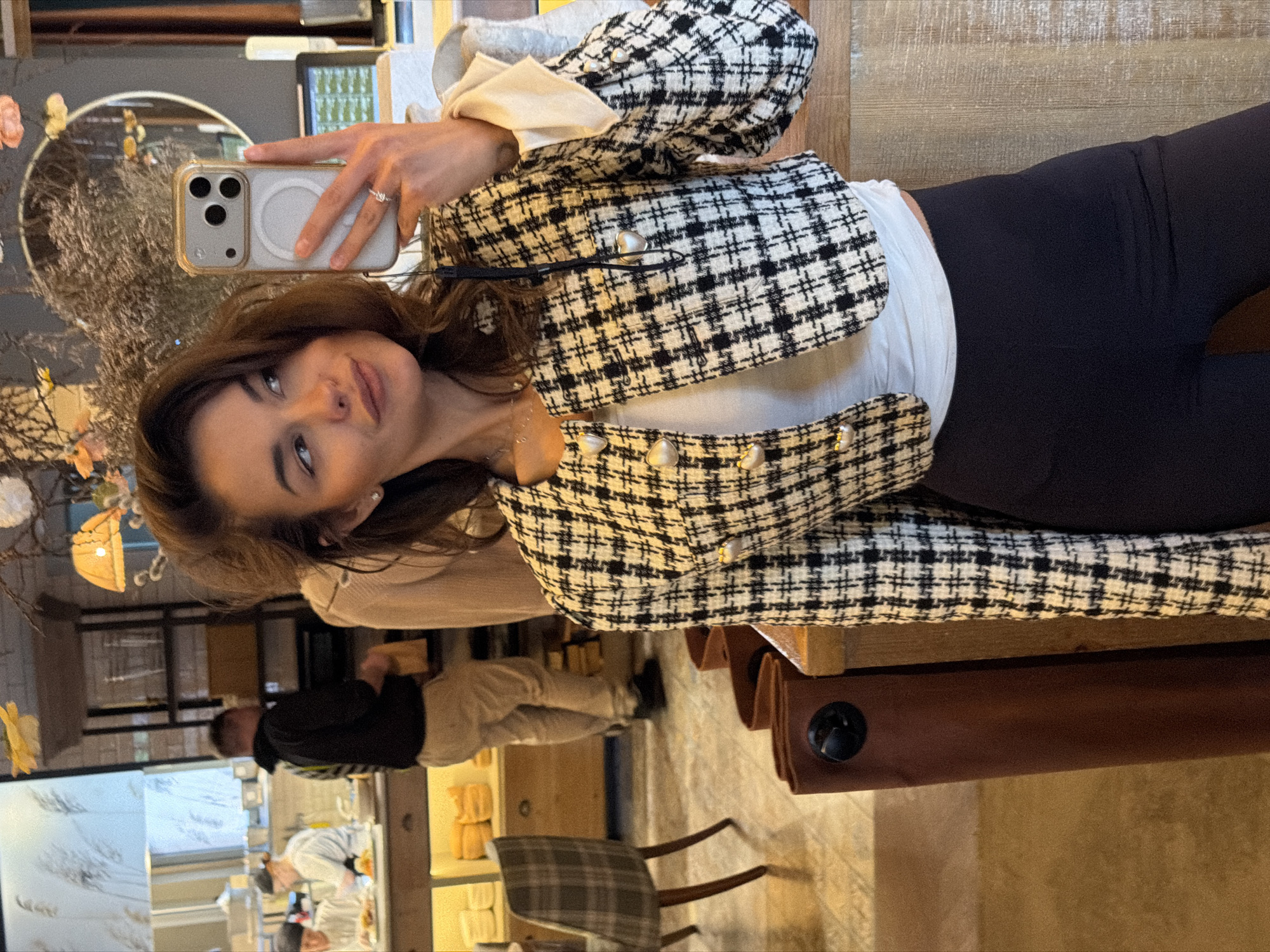 A woman is taking a selfie in an indoor setting. She is wearing a black and white tweed jacket over a white top and black pants. She is holding a smartphone in her left hand. The environment appears to be a cafe or restaurant in Krasnoyarsk, Russia, based on contextual clues such as the presence of cafe furniture, other patrons and staff visible in the background. There is a reflection visible.