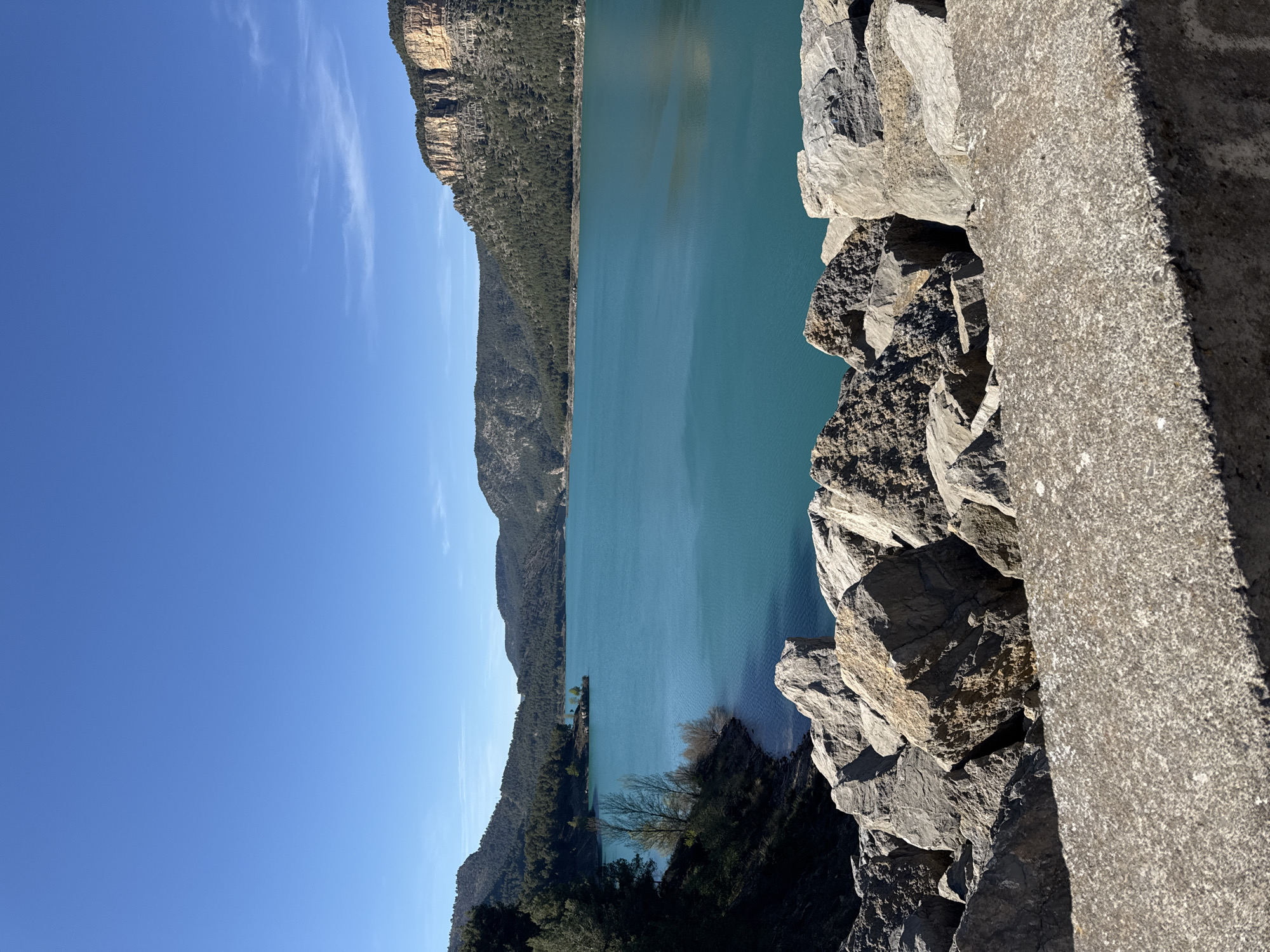 The photograph shows a scenic view overlooking a body of water, likely a lake or reservoir, from what appears to be a cliff or elevated structure. The water is a striking turquoise color. In the background, mountains rise from the opposite side of the water, covered with trees. The sky is a clear blue with wispy clouds near the horizon. In the foreground, rough rocks and a concrete structure border the view.
Based on the visual information and known features of Montanejos, Spain, it is possible that the image depicts the view from a vantage point overlooking the Embalse de Arenoso or the Míjar River area near Montanejos. The turquoise water color is consistent with the mineral content often found in these waters.