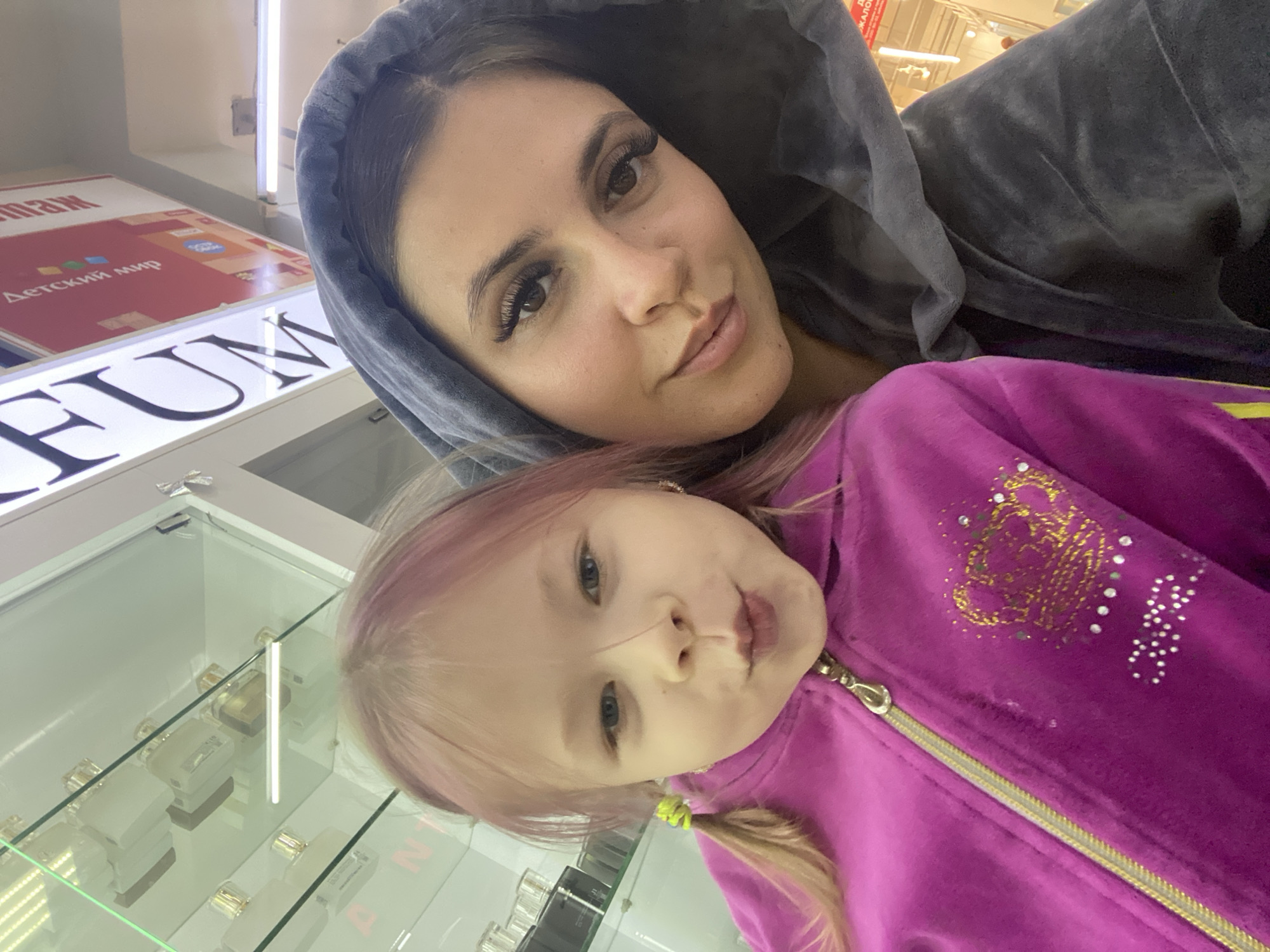 A selfie is taken of a woman and a young child in what appears to be a retail environment, possibly a department store. The woman, with long dark hair and prominent eyelashes, is wearing a grey hooded jacket. The child, with light hair styled into two pigtails and dyed with pink streaks, is wearing a bright pink zippered jacket adorned with a crown and rhinestones. They are posing closely together, with the child making a "pucker" face. In the background, shelves displaying perfume bottles are visible, along with signage that reads "ДЕТСКИЙ МИР" (Children's World) in Russian. The lighting suggests an indoor setting. The presence of "ДЕТСКИЙ МИР" signage indicates the location is a Children's World store, which are common in many Russian cities, including Chaikovsky.