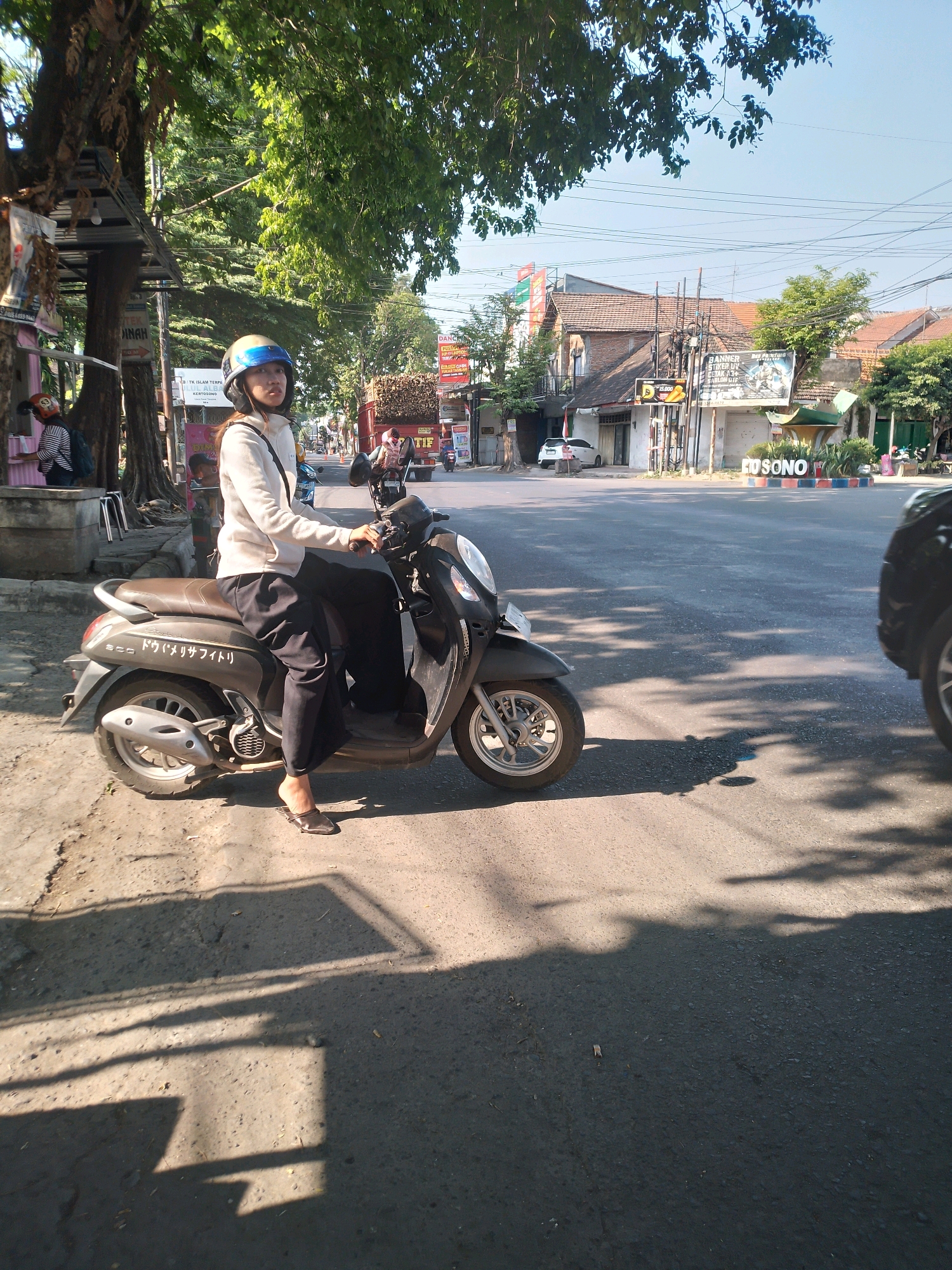 A young woman wearing a helmet and casual clothing is sitting on a dark-colored scooter on a paved road in Kertosono, Indonesia. She is looking forward and appears to be ready to drive. The scooter has Japanese text on its side. In the background, there are buildings, trees, power lines, and other vehicles, suggesting a typical urban street scene. The lighting indicates it is daytime, with shadows cast by trees on the road.