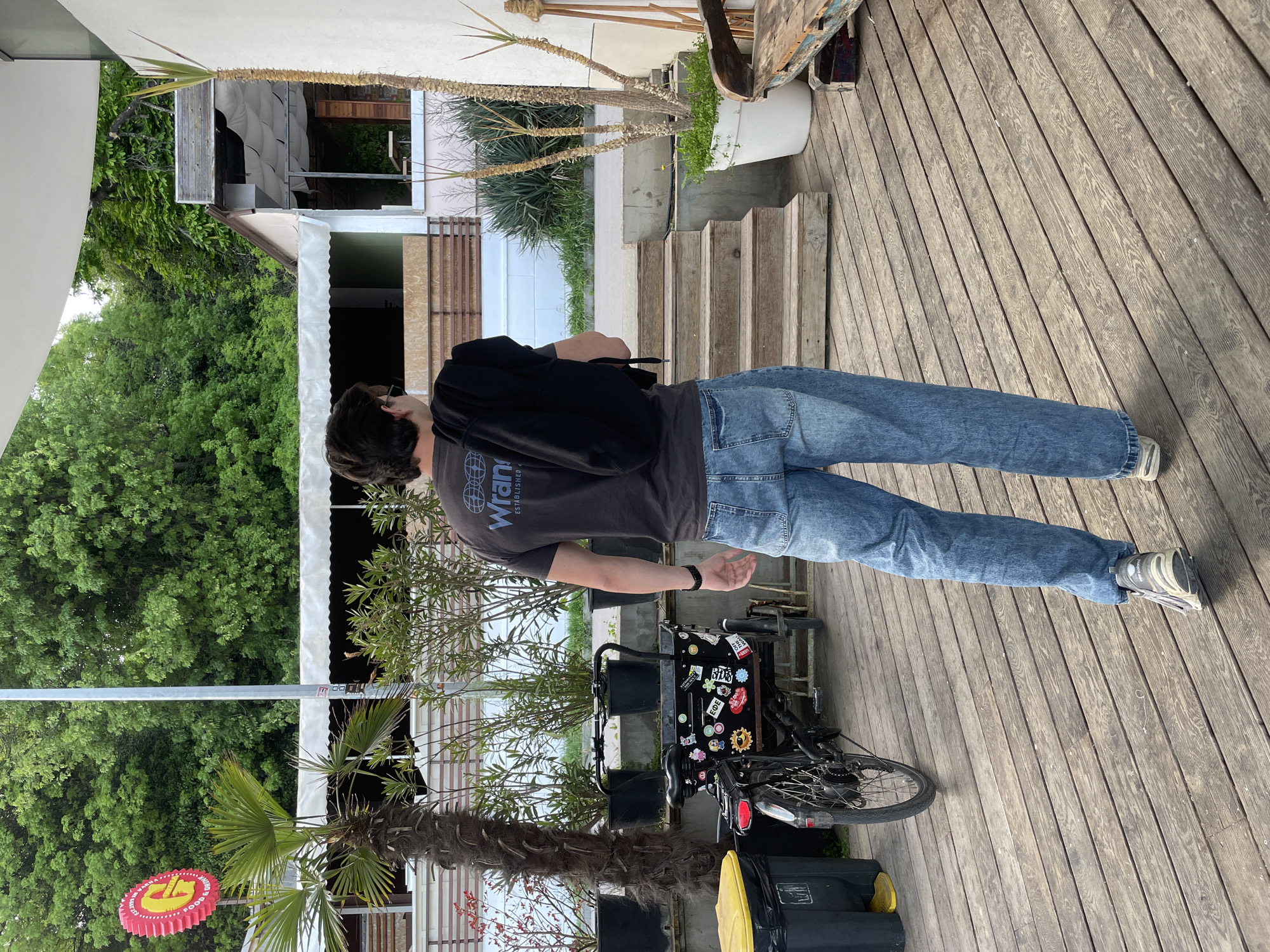 A man with short dark hair, wearing blue jeans, grey t-shirt with a "Wrangler" logo, a black backpack, and grey sneakers, stands on a wooden deck next to a black bicycle. The bicycle has many stickers attached to its frame and a black container attached to the back of it. A palm tree, along with other greenery, flanks the left side of the frame. In the background, there is a building and a set of stairs leading upwards. The city of Varna, Bulgaria is not explicitly evident in the image, so I cannot verify its location.