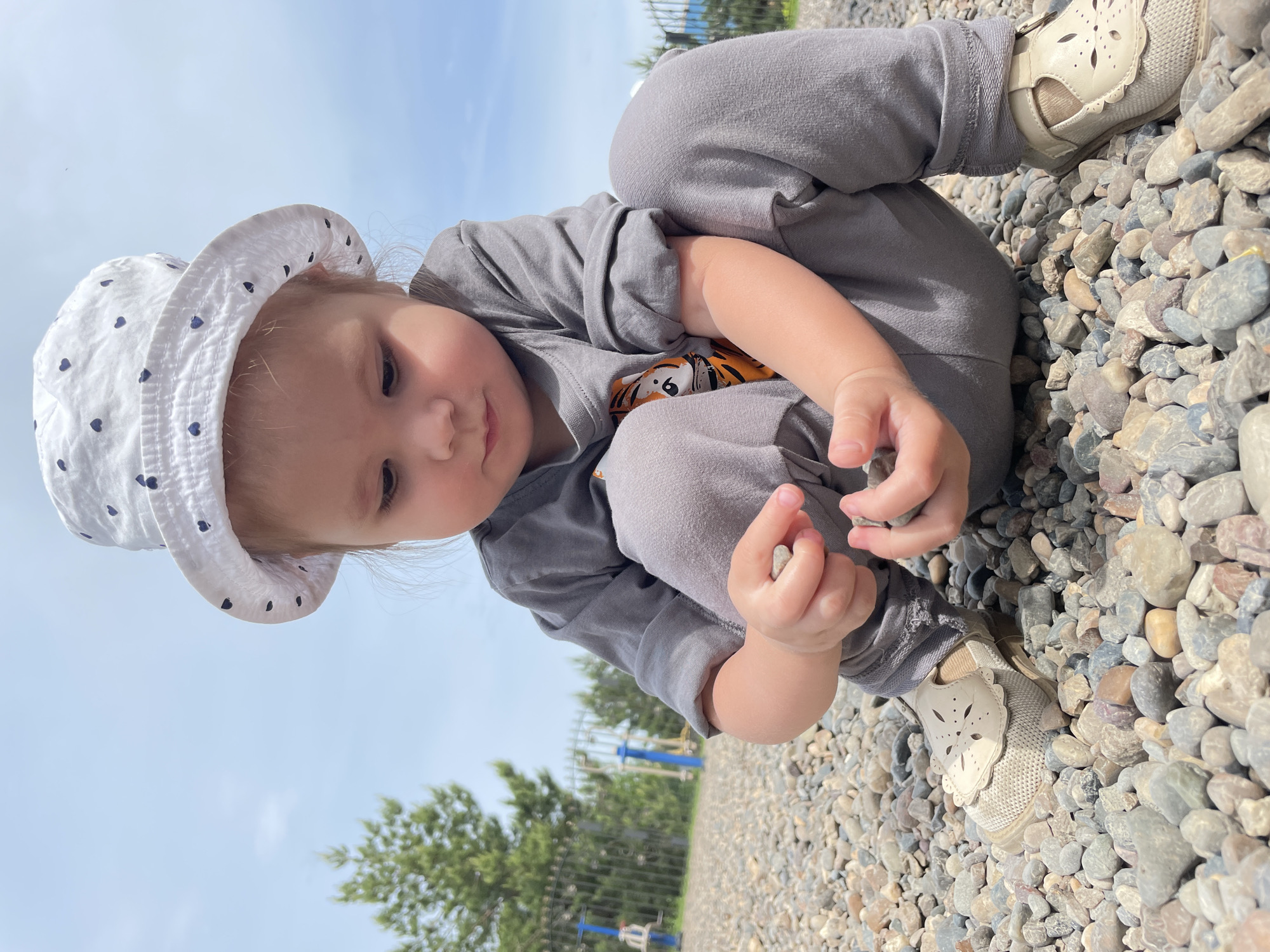 A young child, wearing a grey outfit and a white hat with blue hearts, is sitting on a ground covered with small rocks. The child is holding a rock in their hand. The child is wearing beige shoes. In the background, there are green trees and a blue sky. The location is a playground in Izhevsk, Russia.