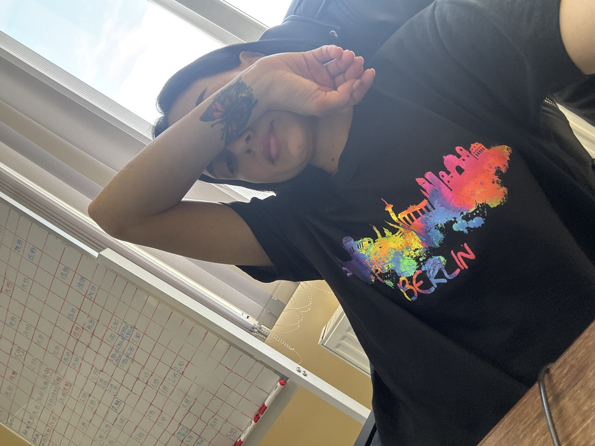 A person wearing a black t-shirt with a colorful "BERLIN" print on it is partially obscuring their face with their hand, which has a butterfly tattoo. The person appears to be indoors, with window blinds visible in the background and a white board with what seems to be handwritten financial data on it. The environment includes hints of window and wall. The person is positioned at an angle, looking towards the camera. The image does not contain any discernible information about Tbilisi, Georgia.